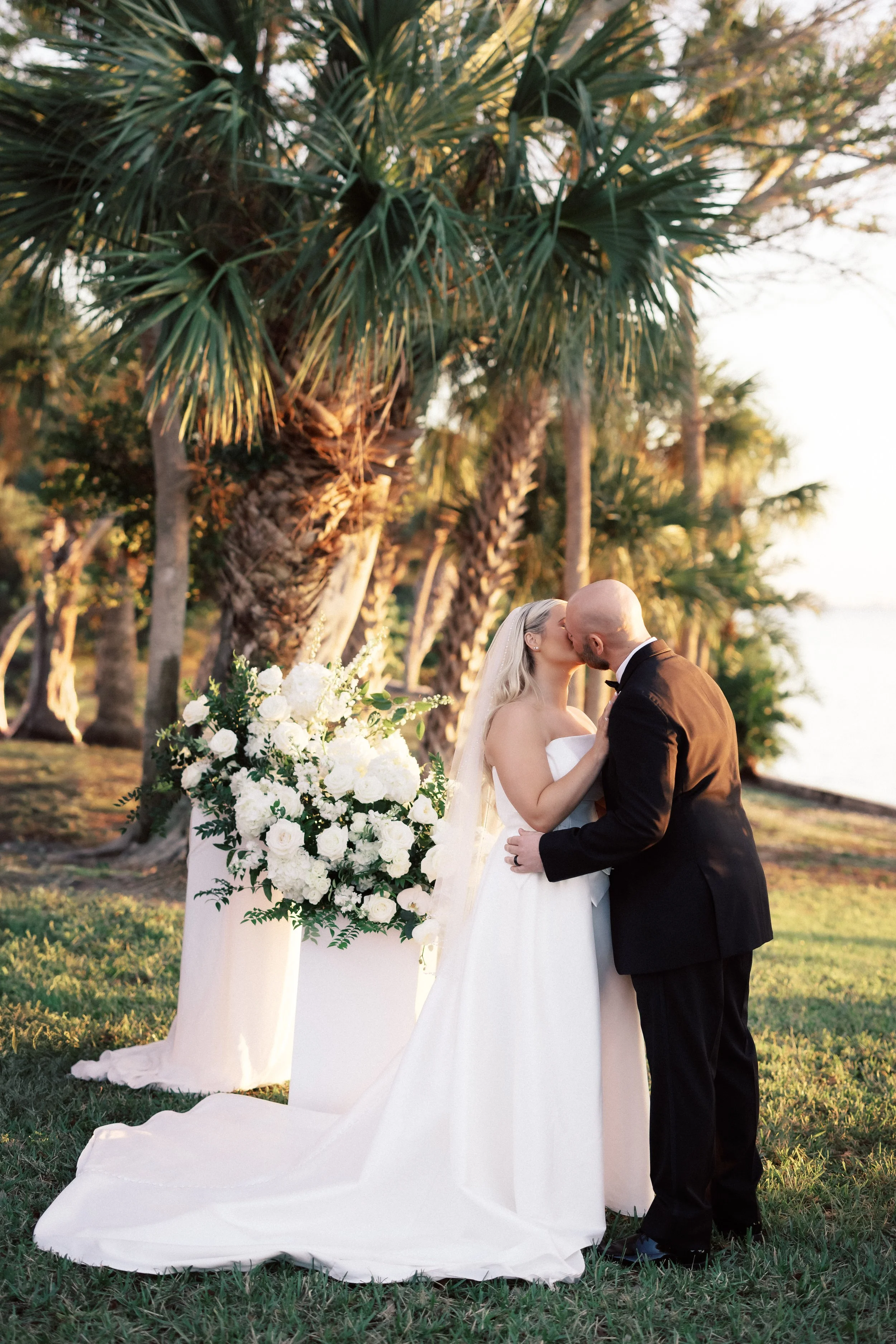 A bride and groom kissing during their outdoor wedding ceremony near palm trees and water. The bride is wearing a white strapless wedding gown with a long train, and the groom is in a black tuxedo. There are white floral arrangements on a table nearb
