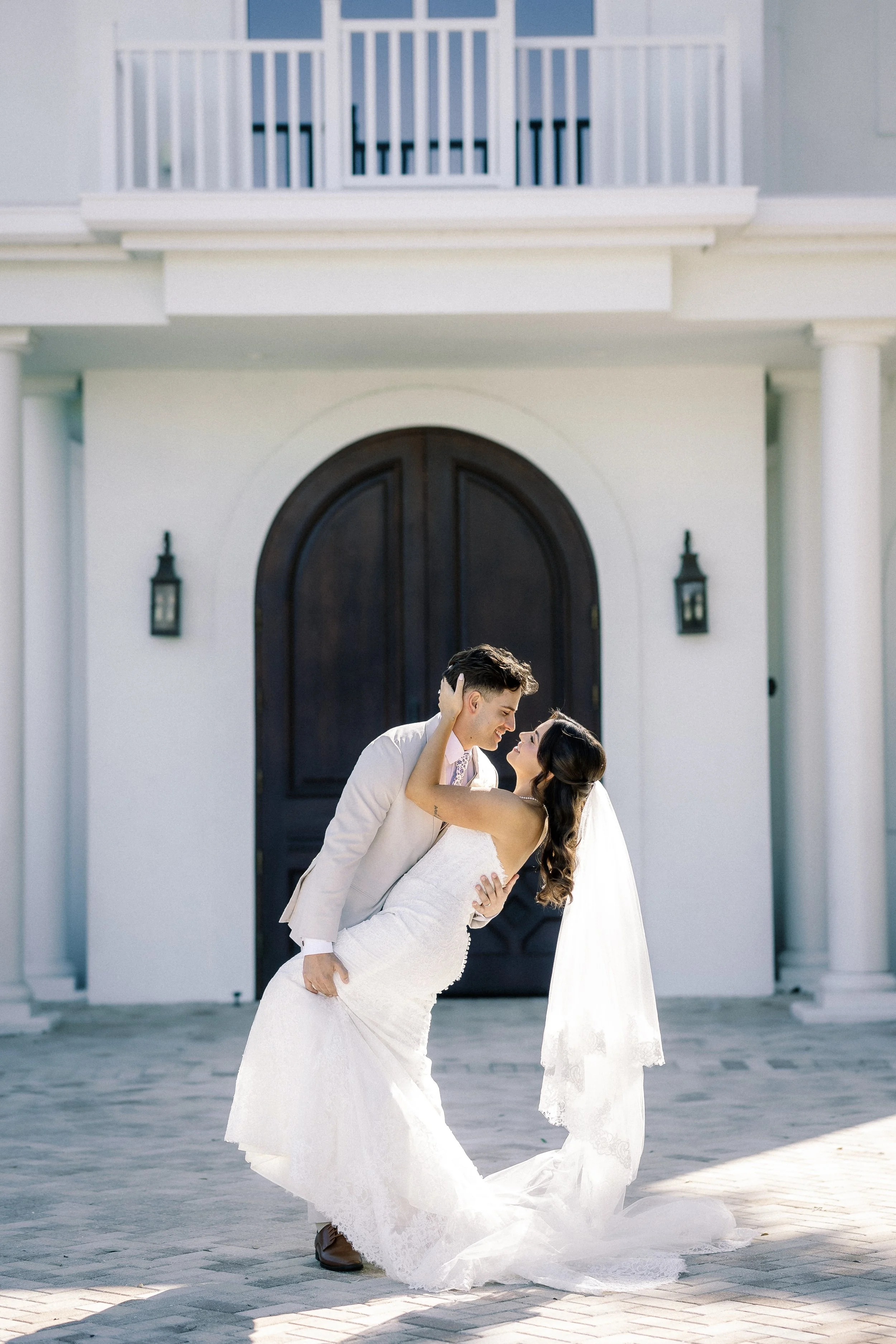 A bride and groom sharing a dance outside a white house with dark front door, columns, and lanterns, on a sunny day.