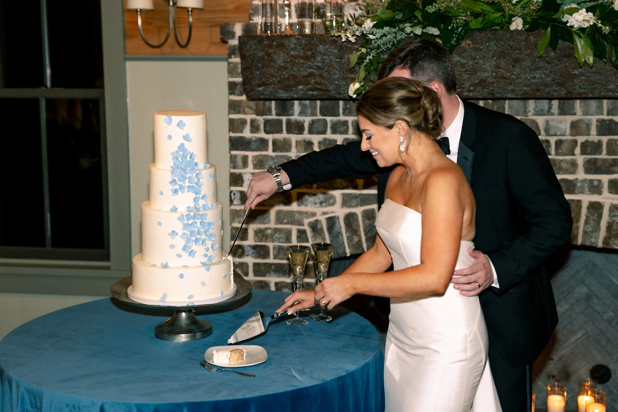 A bride and groom cutting their wedding cake, which is a tall, four-tiered cake decorated with blue flowers, at a wedding reception.