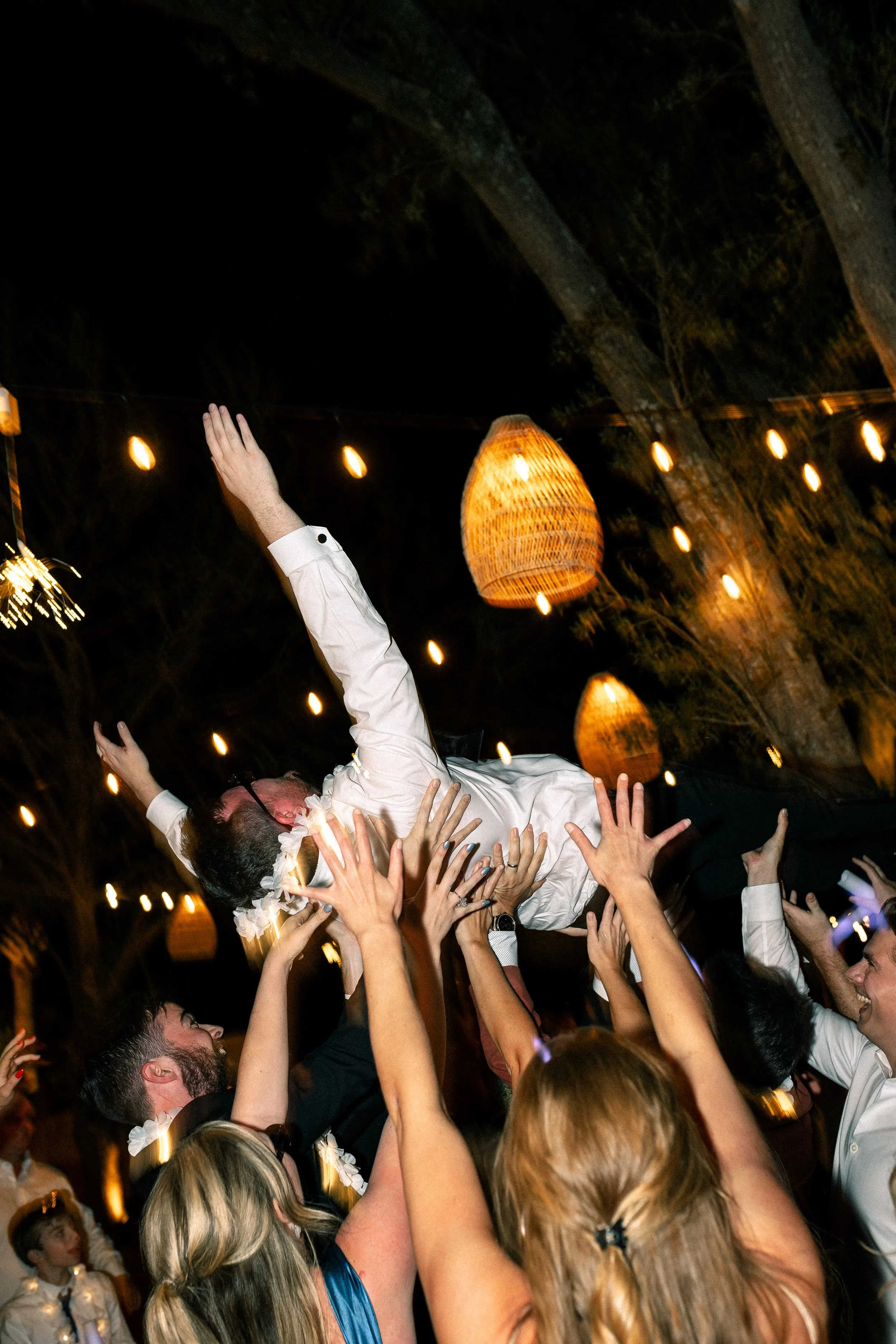 People celebrating at a wedding reception, lifting a groom into the air among string lights and lanterns at night.