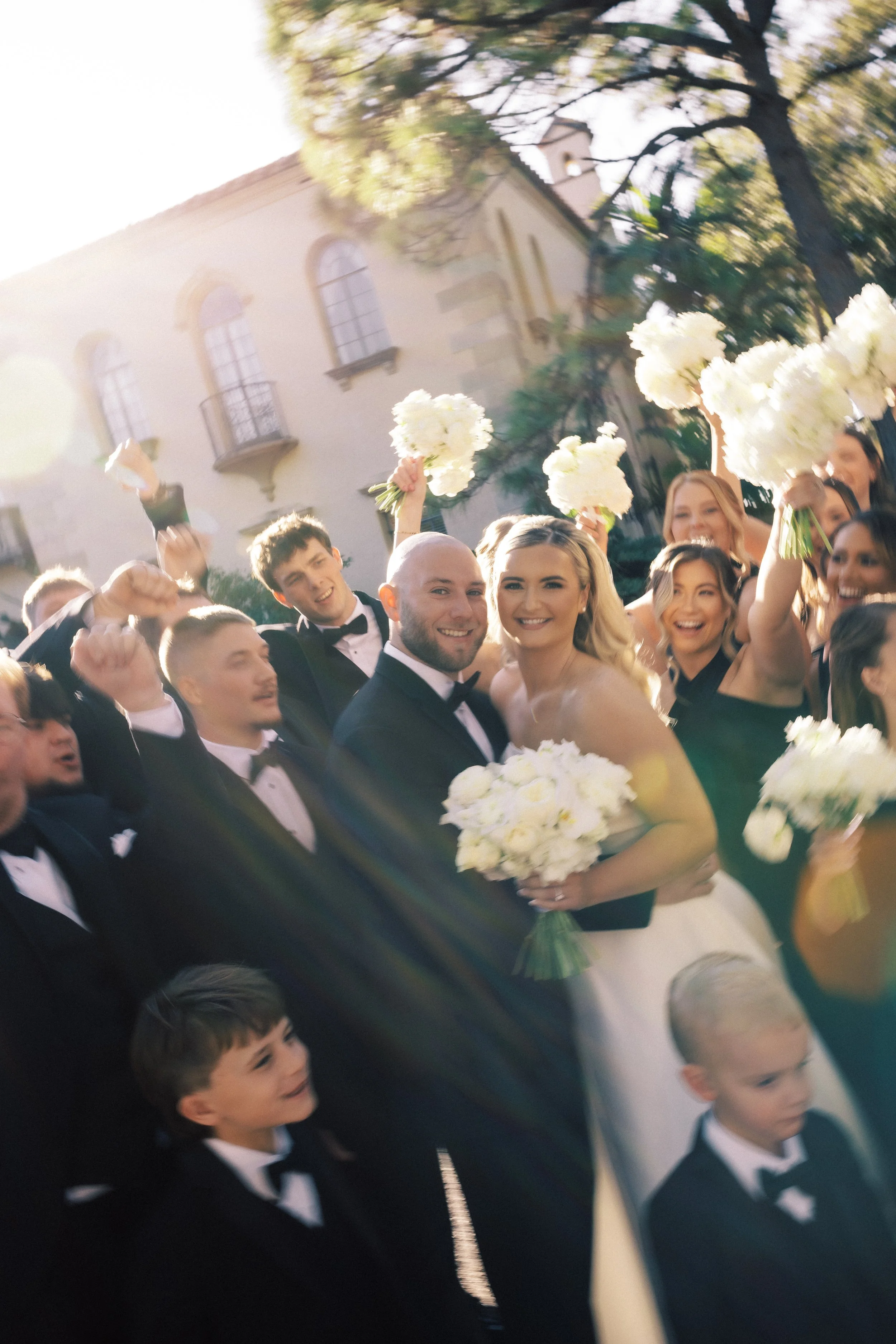 A wedding celebration outdoors with the bride and groom surrounded by friends and family, many of whom are holding bouquets of white flowers, in front of a building with arched windows and surrounded by trees.