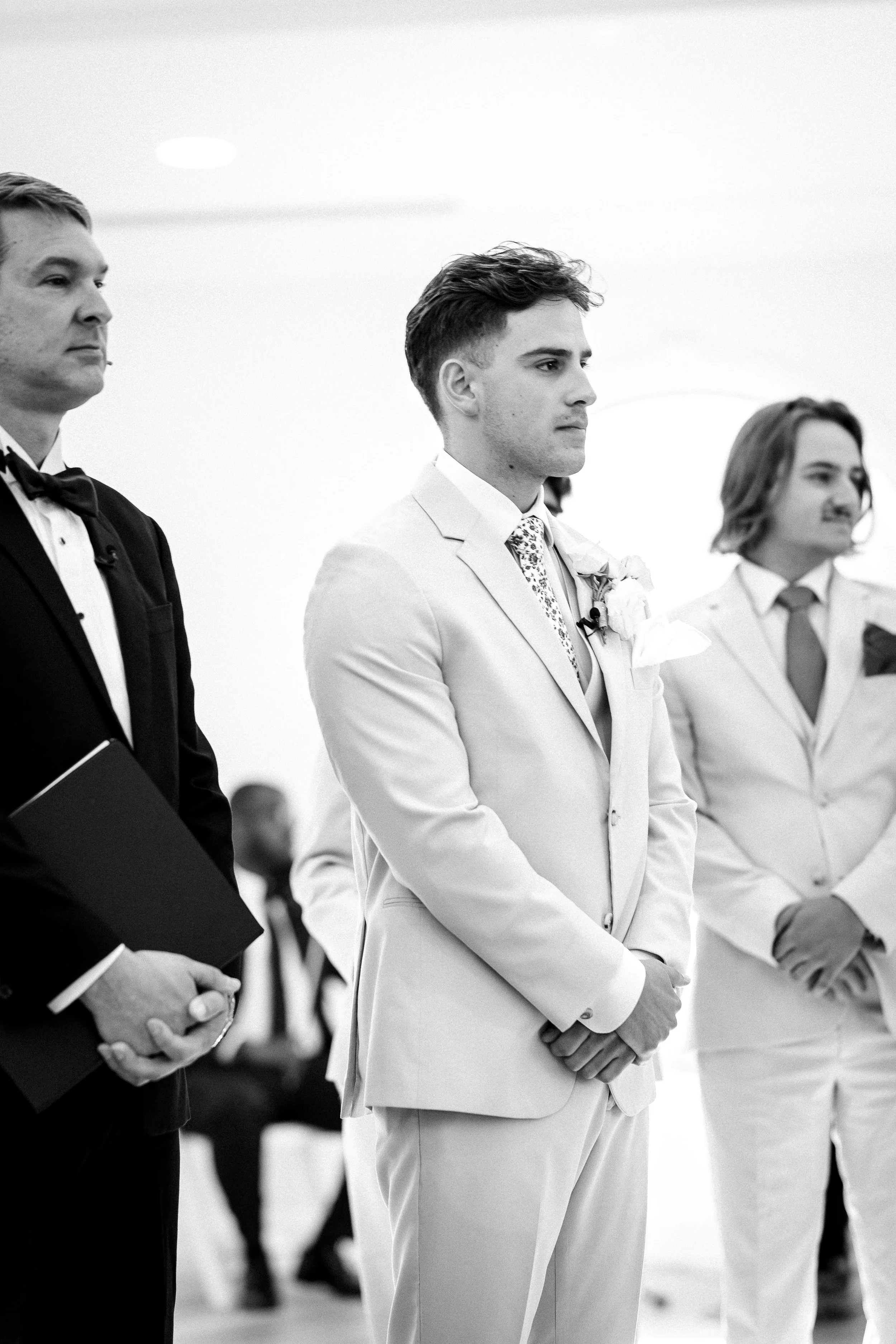 A black and white photo of a wedding ceremony showing a groom in a light-colored suit standing with his hands clasped, flanked by two groomsmen dressed in dark and light suits, with some guests blurred in the background.