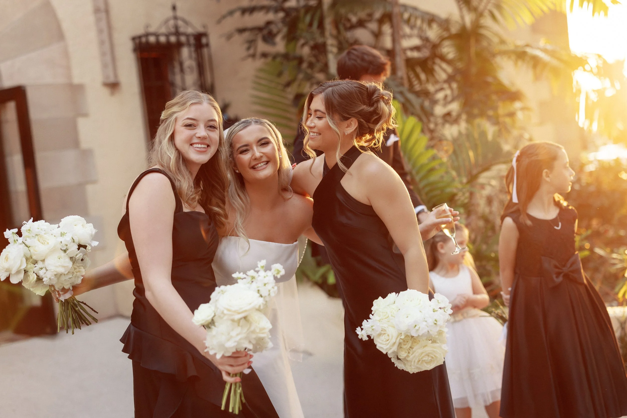 Three women smiling and holding bouquets of white flowers at a wedding reception outdoors during sunset, with other guests in the background.