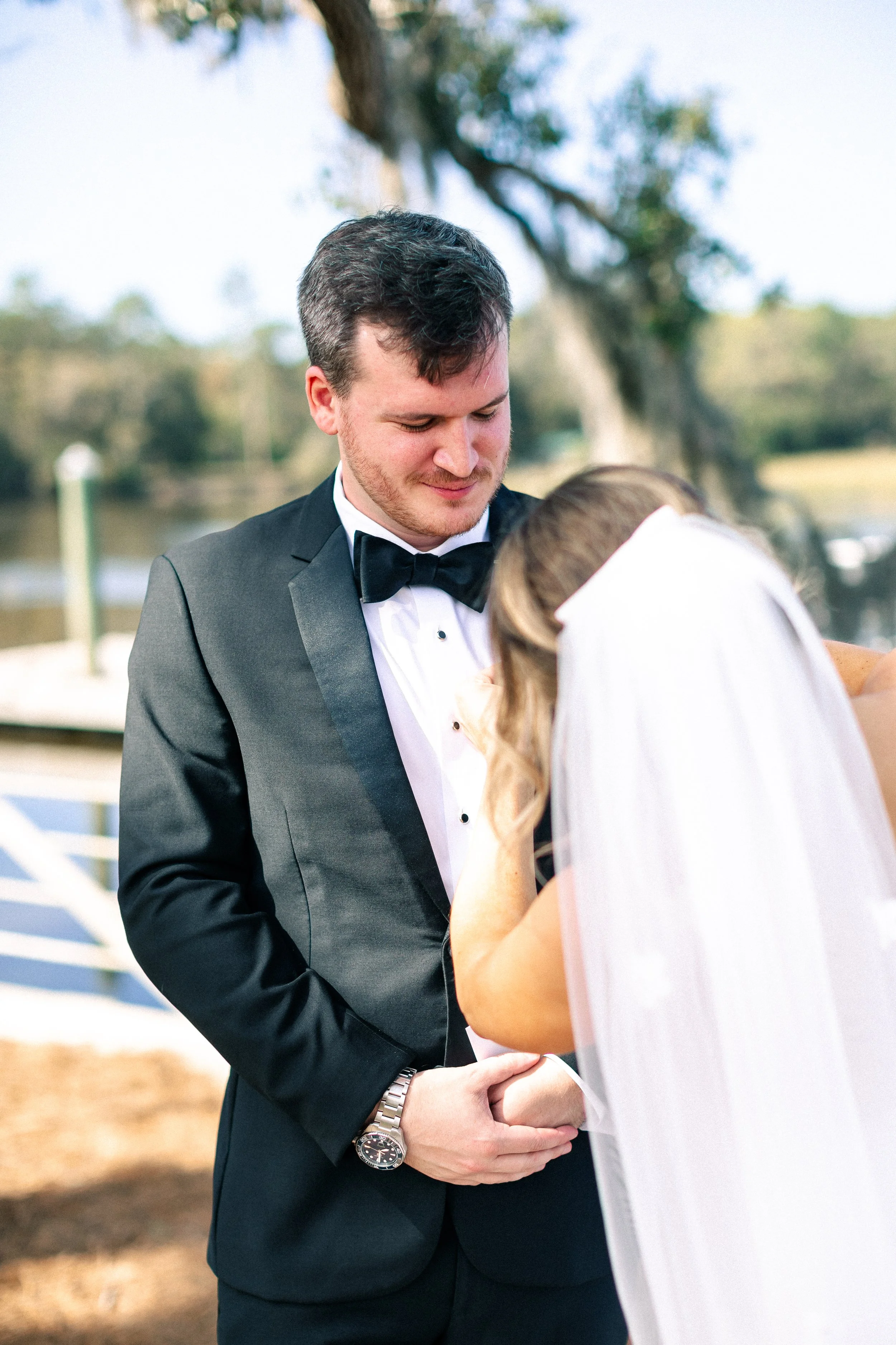 A bride and groom during their wedding ceremony outdoors, with the groom wearing a black tuxedo and the bride in a white wedding dress and veil, holding hands and looking down.