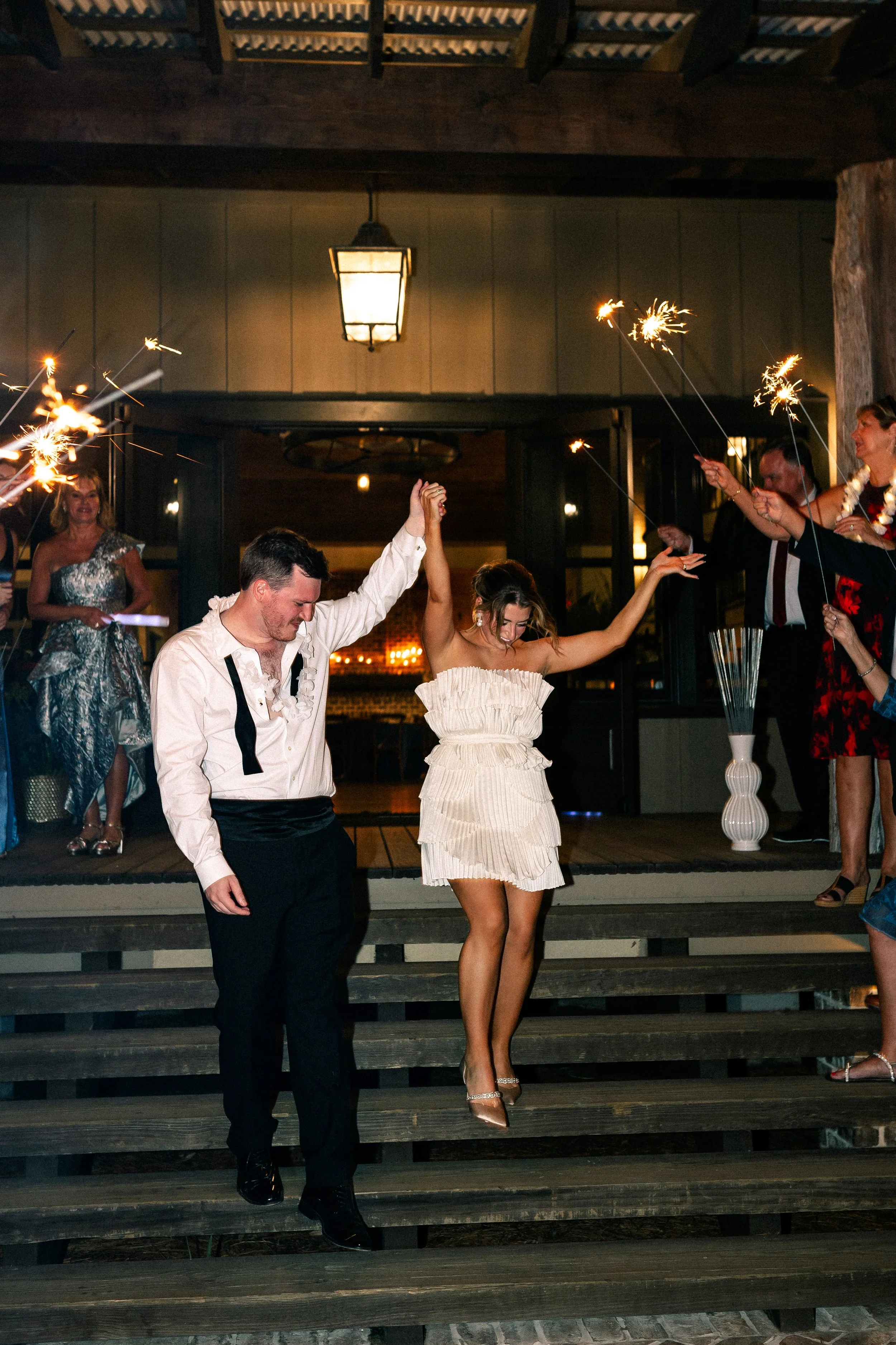A newlywed couple holding hands and dancing down the steps with sparklers in a celebration at night.