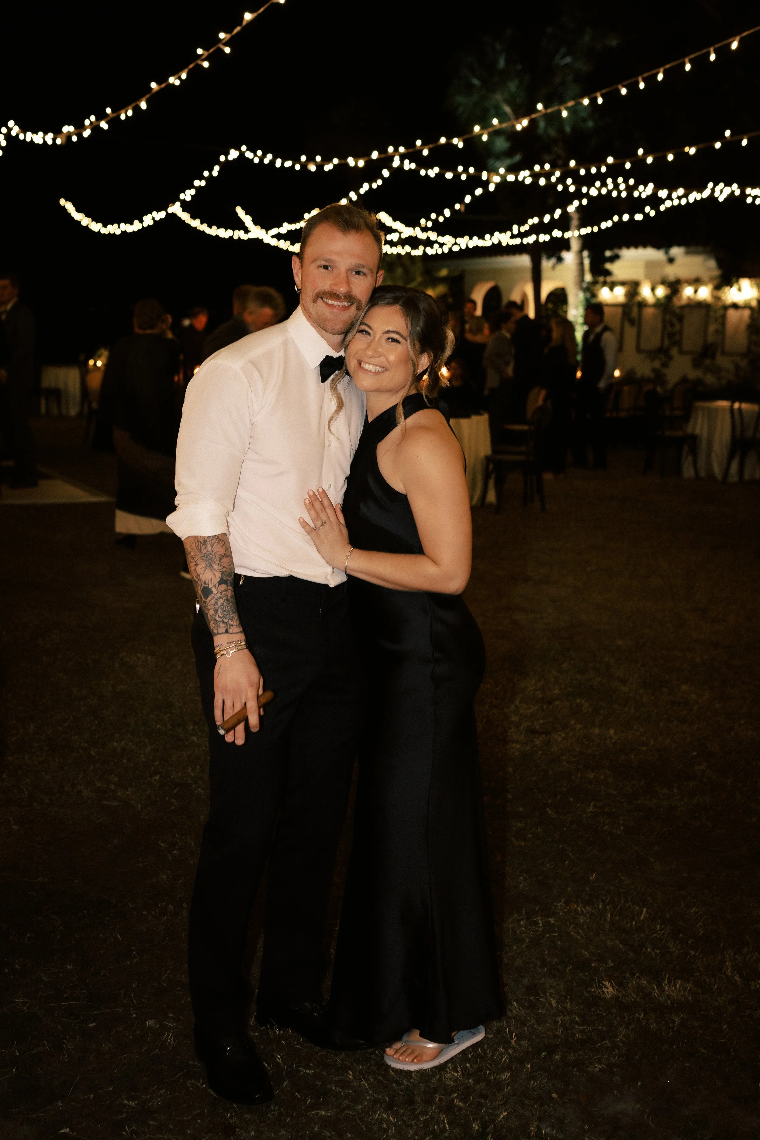 A couple dressed elegantly at a nighttime outdoor event, with string lights overhead, smiling and hugging.