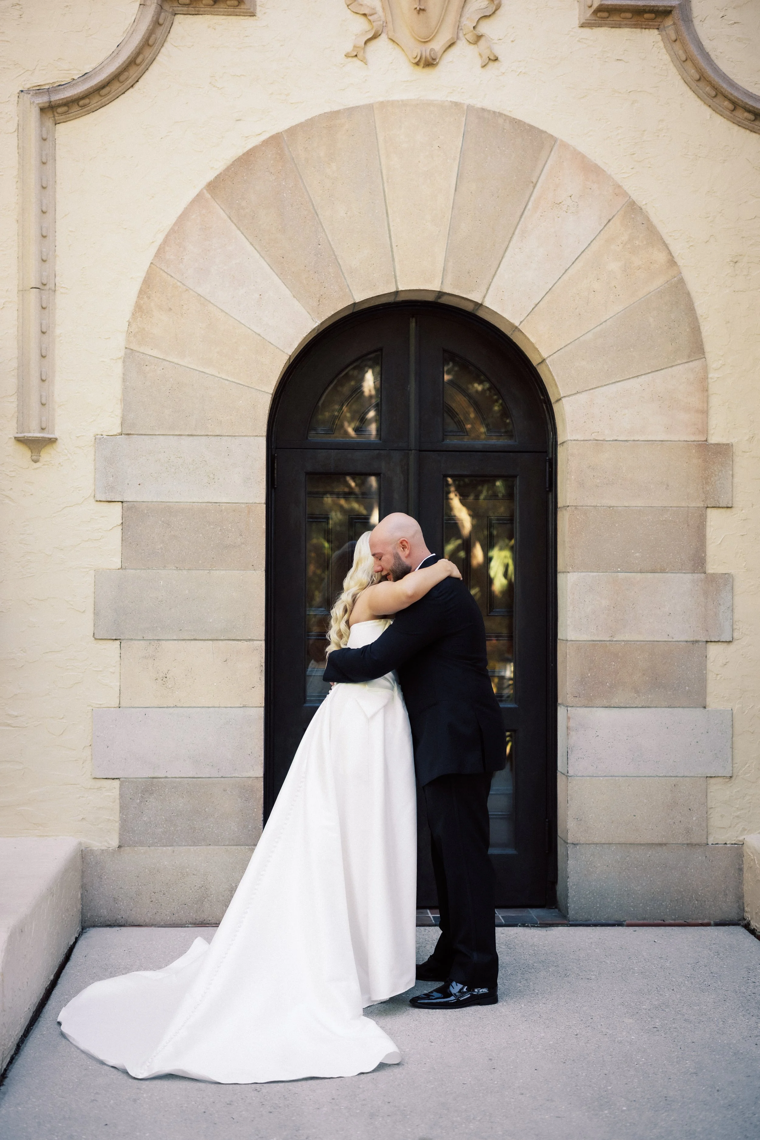 A bride and groom embrace each other outside a building with a large arched window and beige stone and stucco wall.