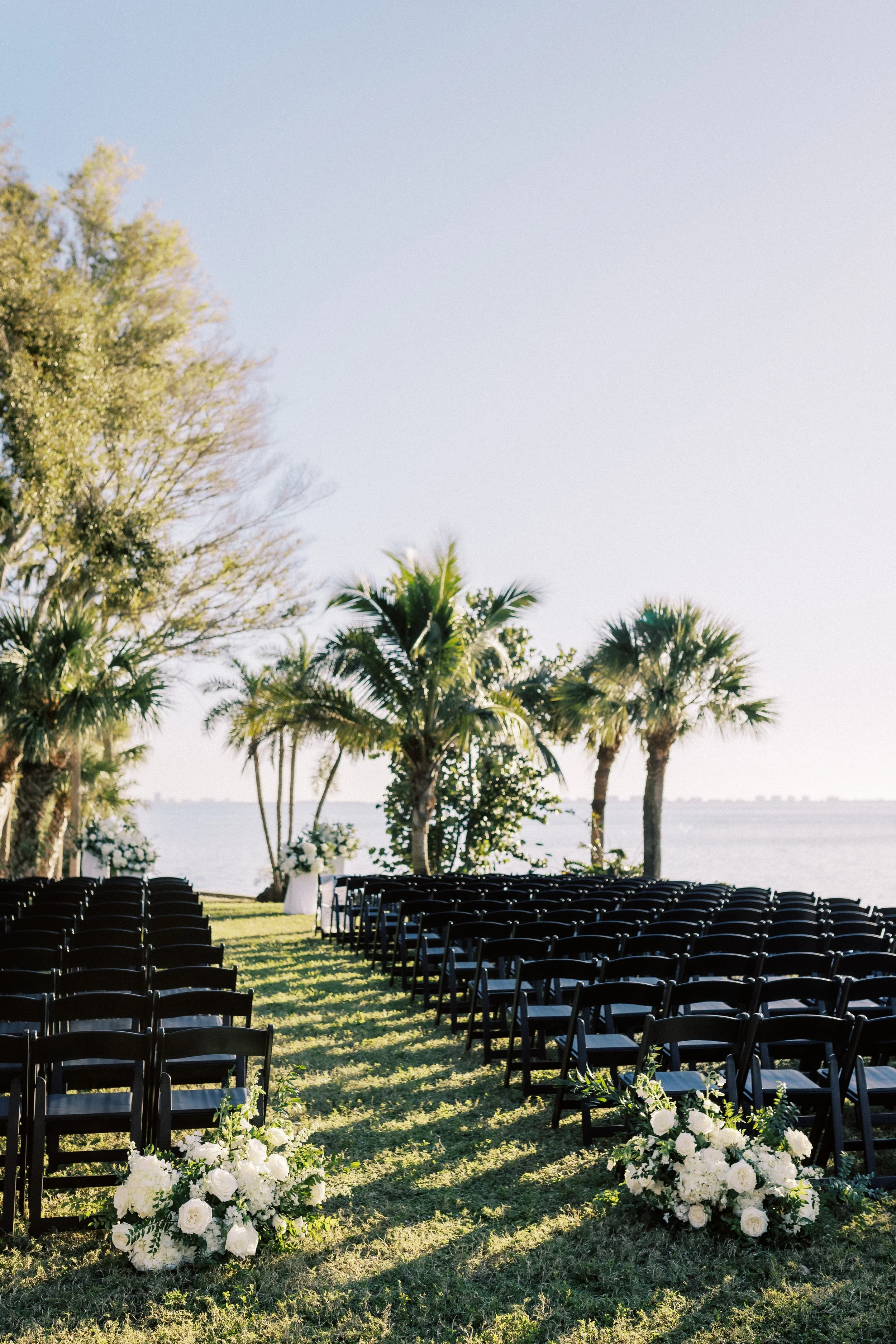 Outdoor wedding setup with chairs, floral arrangements, palm trees, and water in the background during daytime.
