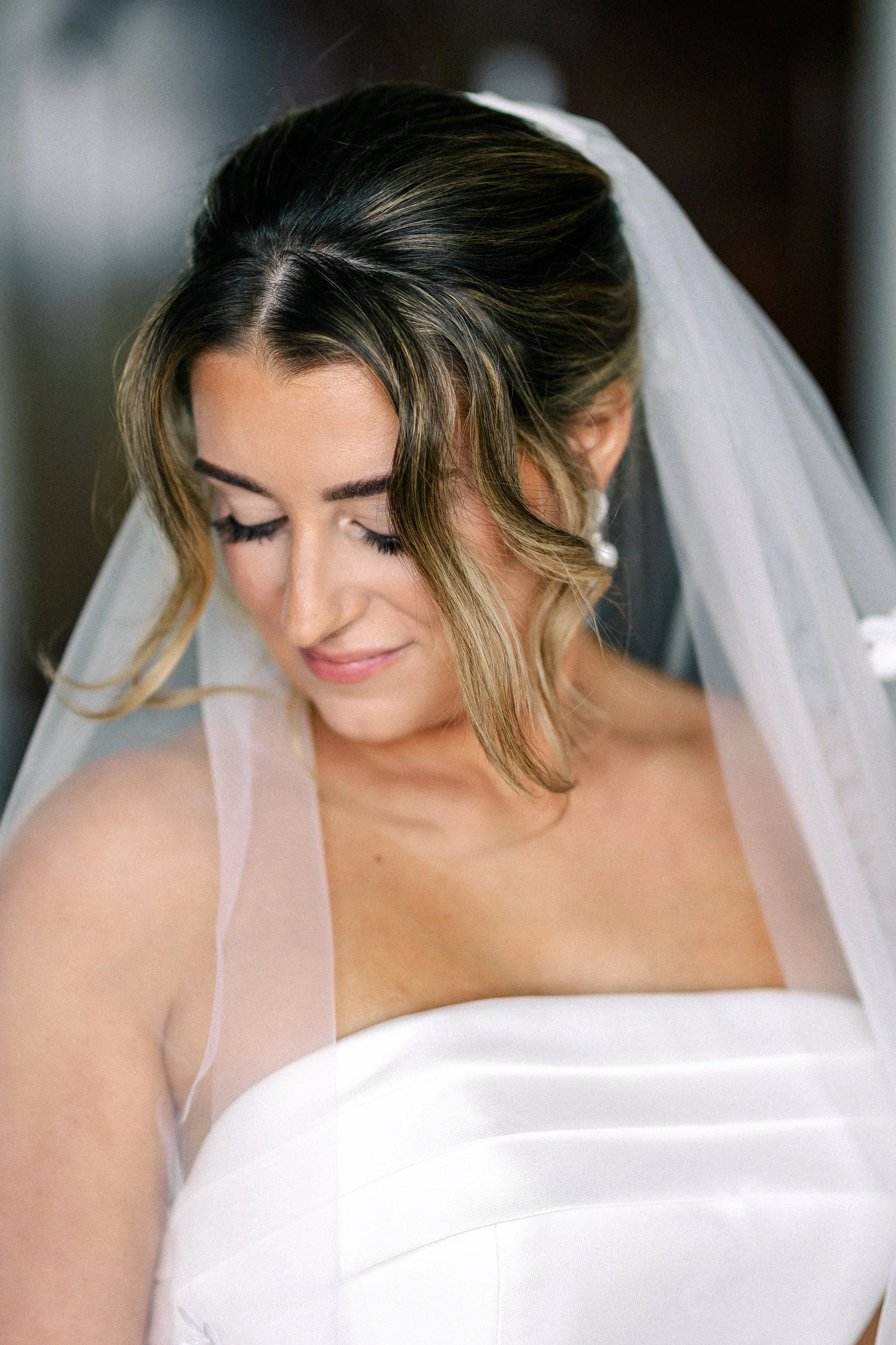 Close-up of a bride with styled hair and veil, wearing a strapless white wedding dress, looking down with a gentle smile.