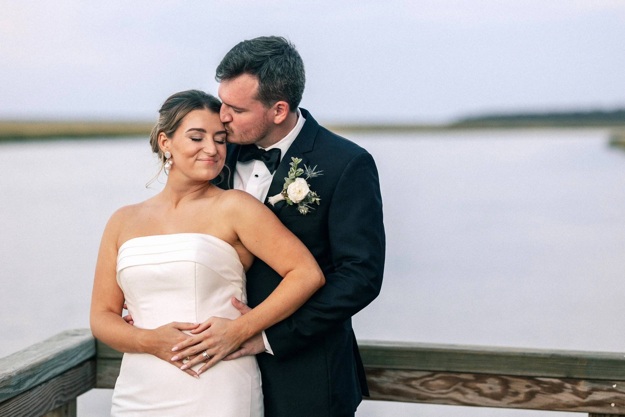 A newlywed couple standing on a wooden dock by a body of water, with the groom kissing the bride on her forehead, both smiling with eyes closed.