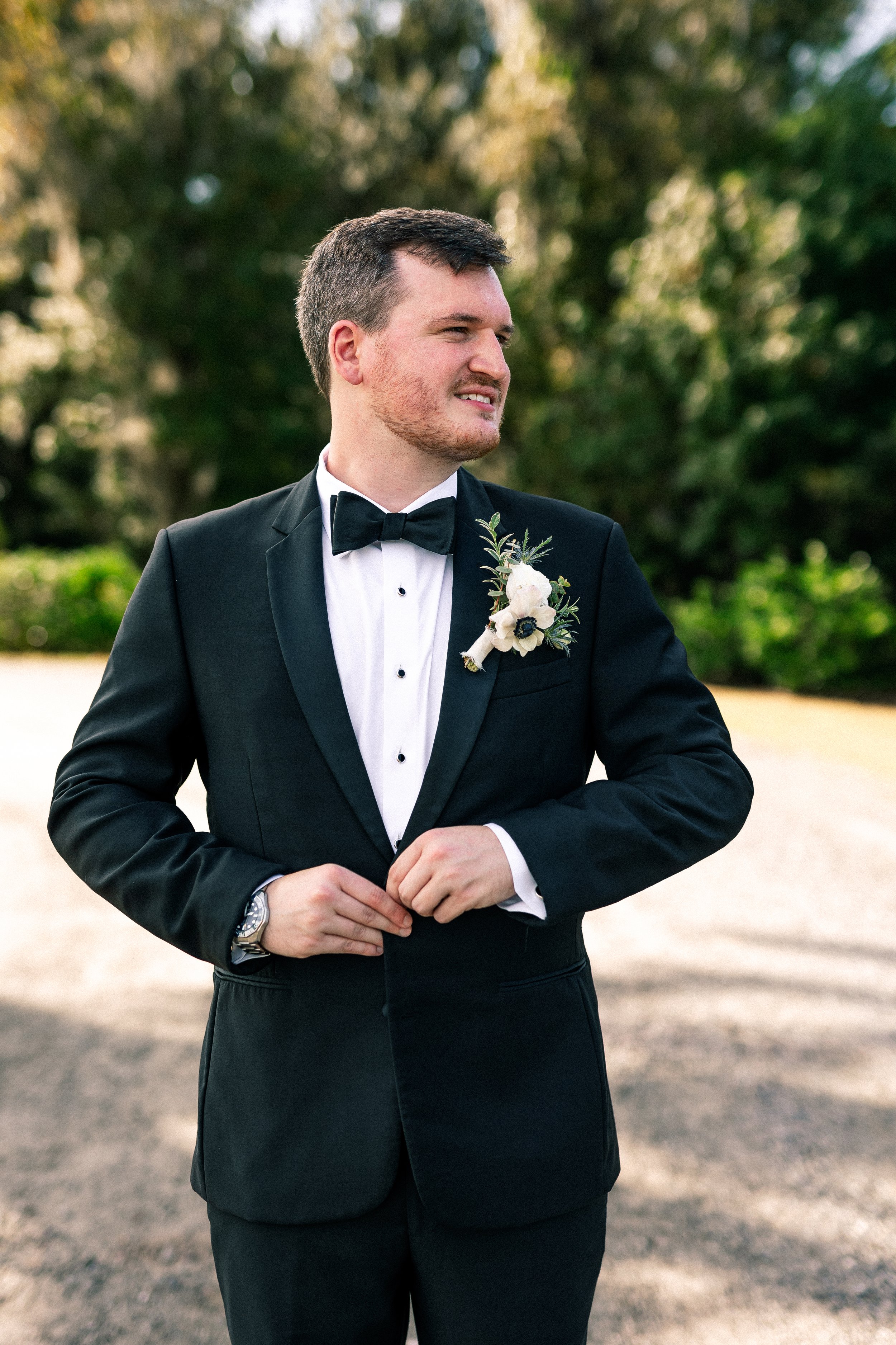 A young man in a black tuxedo with a white shirt and black bow tie, standing outdoors with trees in the background. He is adjusting his jacket, wearing a wristwatch, and has a boutonniere with white and dark flowers pinned to his lapel. He is smiling