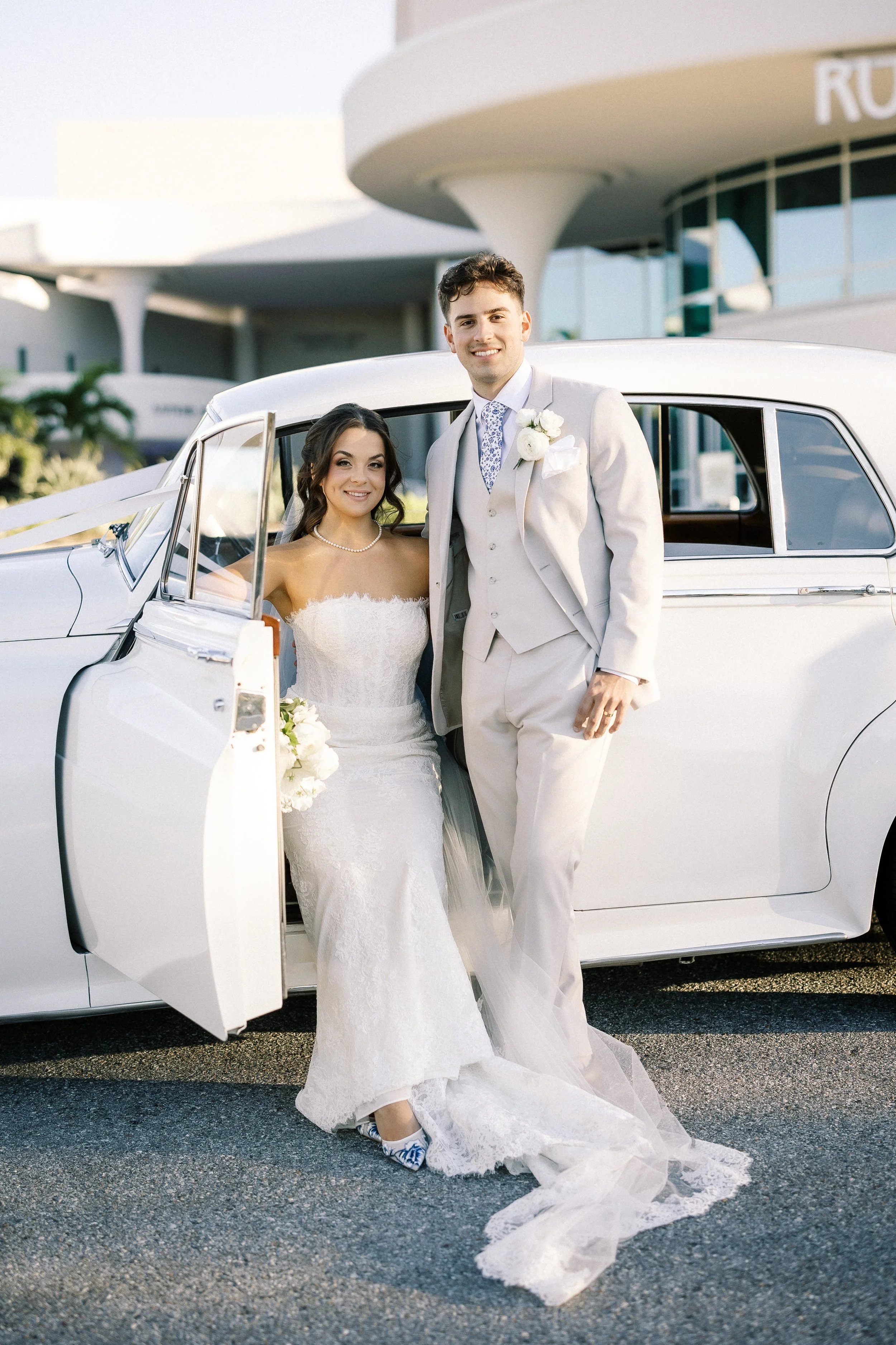 A bride in a white wedding dress and a groom in a light-colored suit standing outside next to a classic white car, smiling.