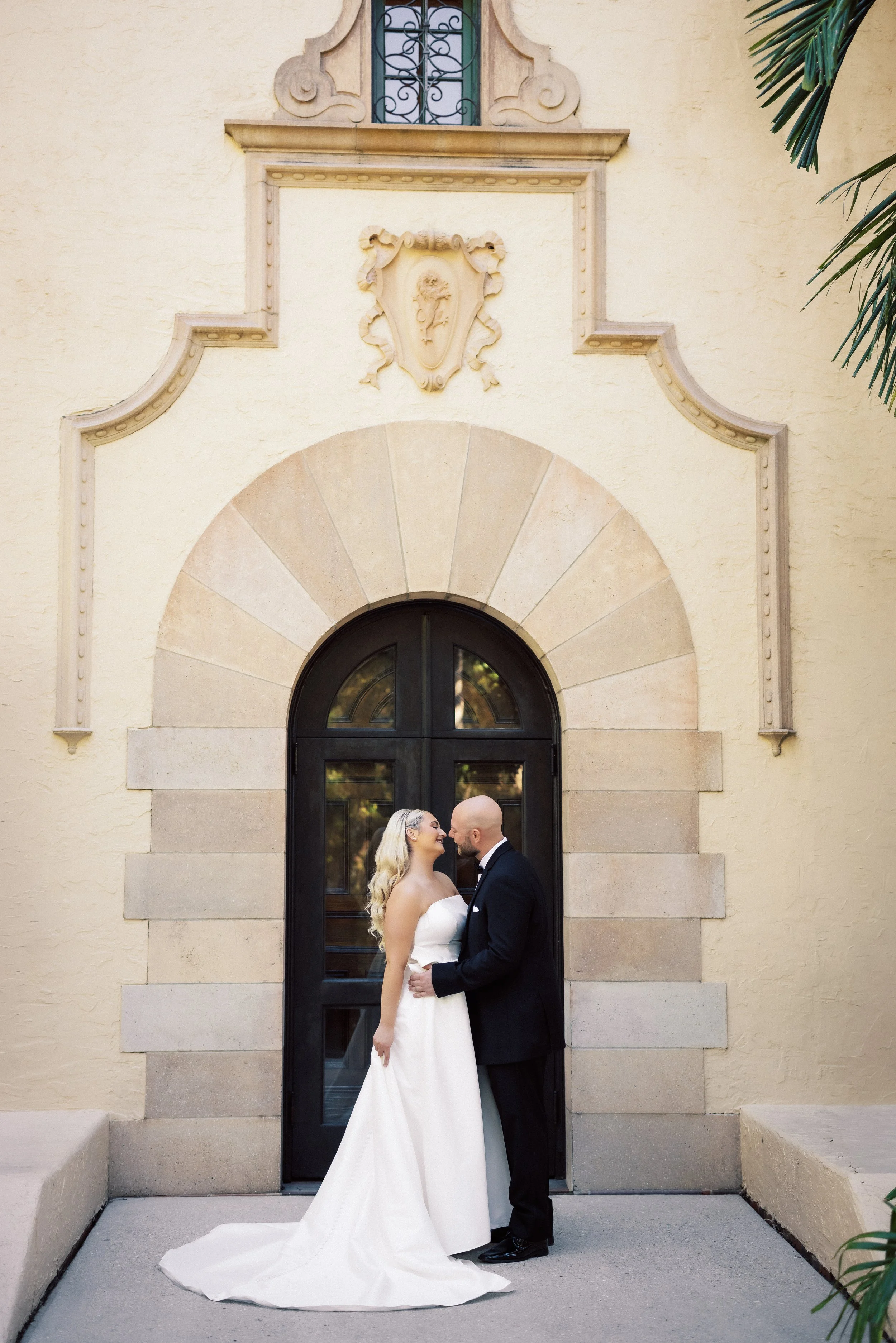 Bride and groom share a kiss in front of a historic building with an arched door and decorative stone trim.