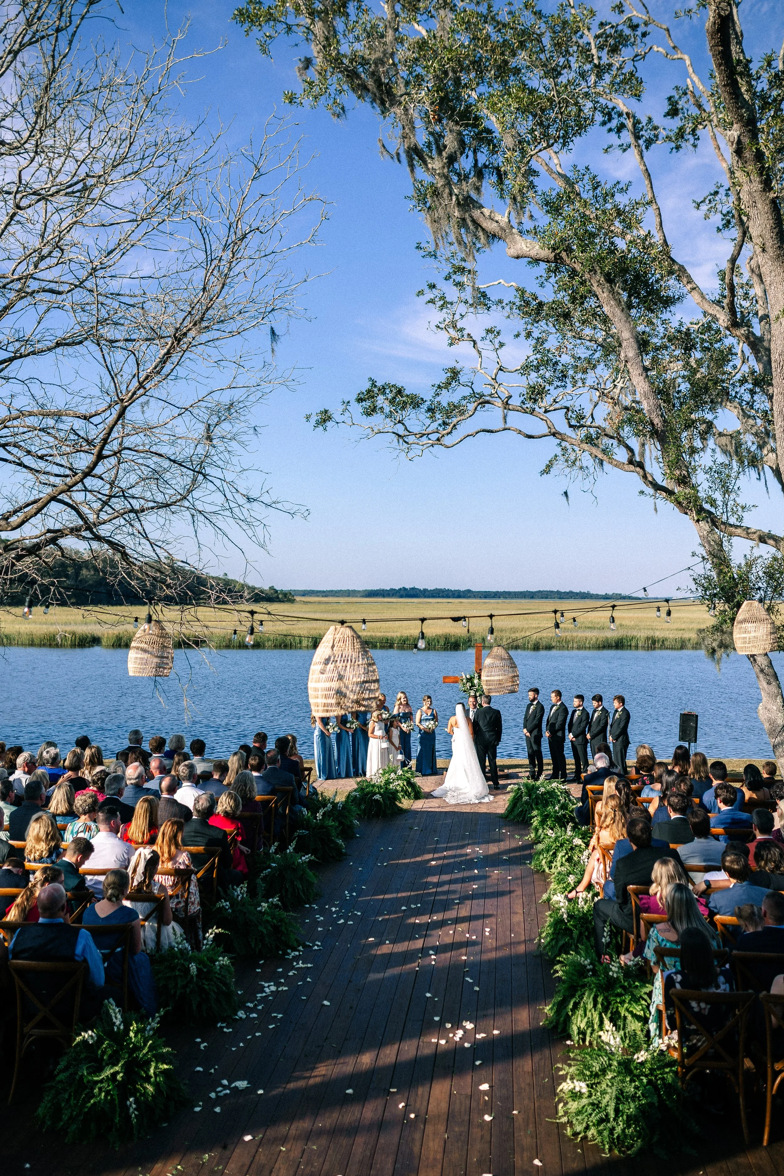 An outdoor wedding ceremony by a lake with guests seated on wooden chairs, a couple standing at the altar with a officiant, surrounded by greenery and hanging wicker lanterns, under a clear blue sky.