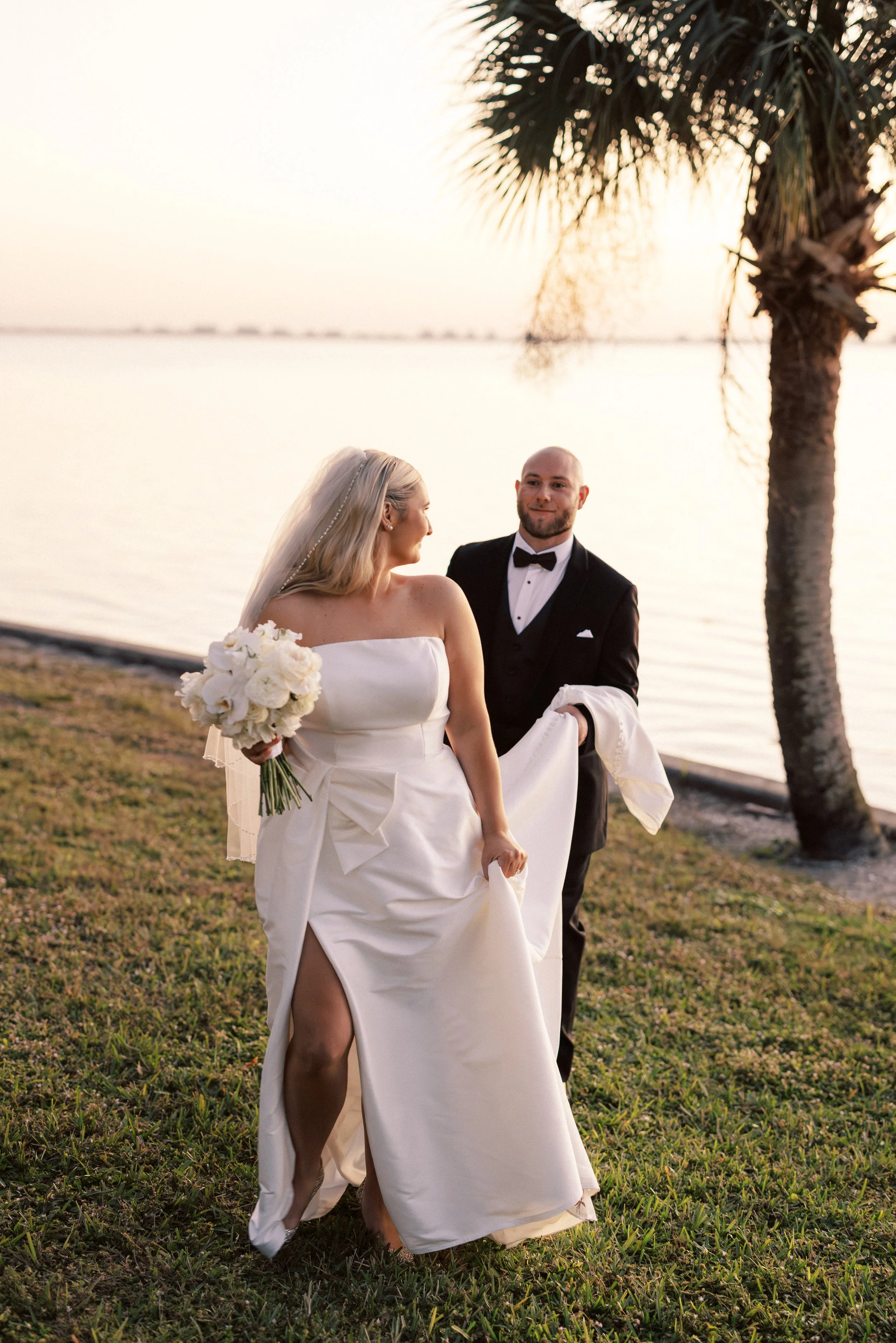 Bride and groom on a grassy lakeside at sunset, with a palm tree nearby. The bride is holding a bouquet and lifting her wedding dress while the groom stands in a tuxedo, looking at her.