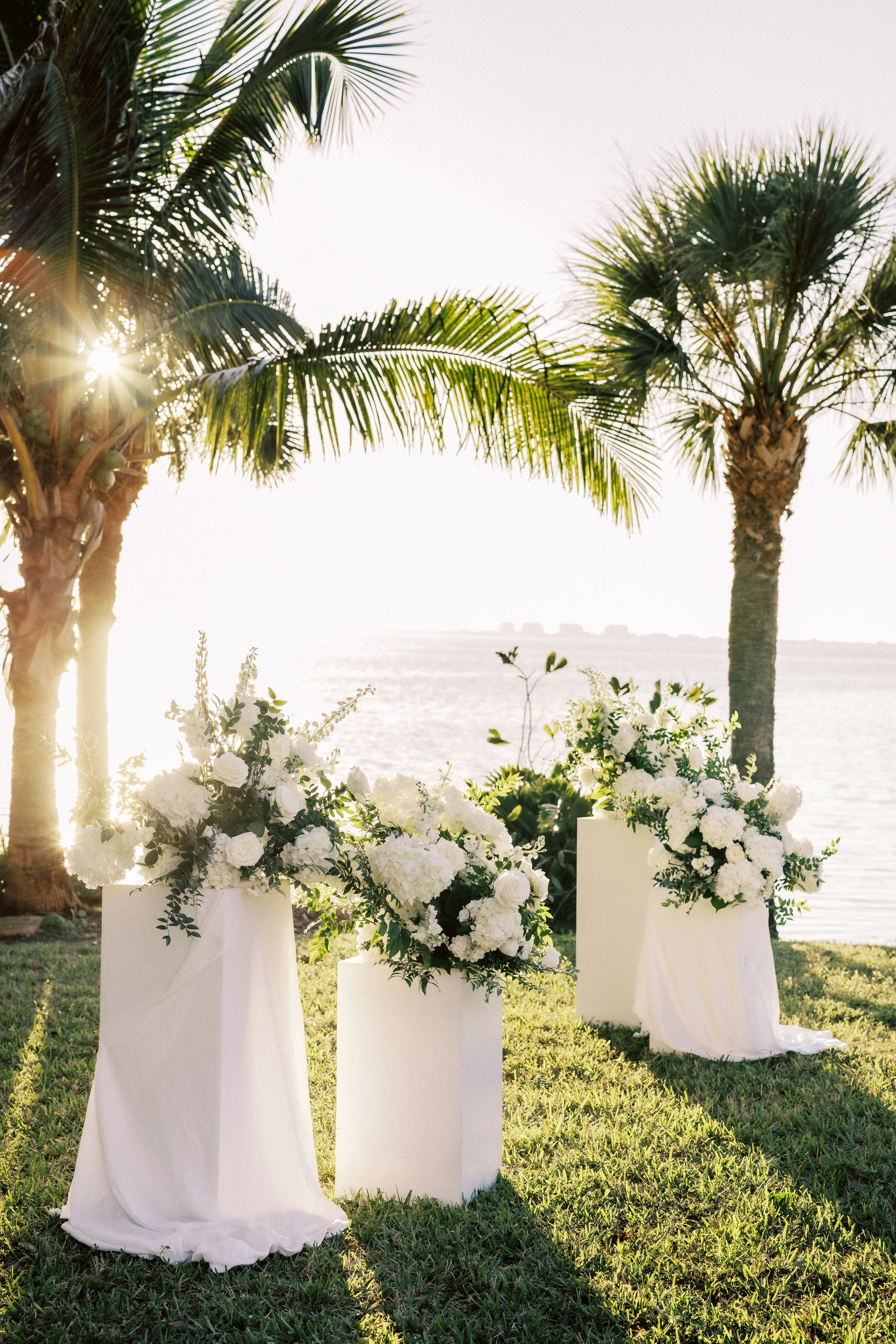 Tropical outdoor wedding altar with white floral arrangements on tall white stands, set by a lake or ocean with palm trees and sunlight in the background.