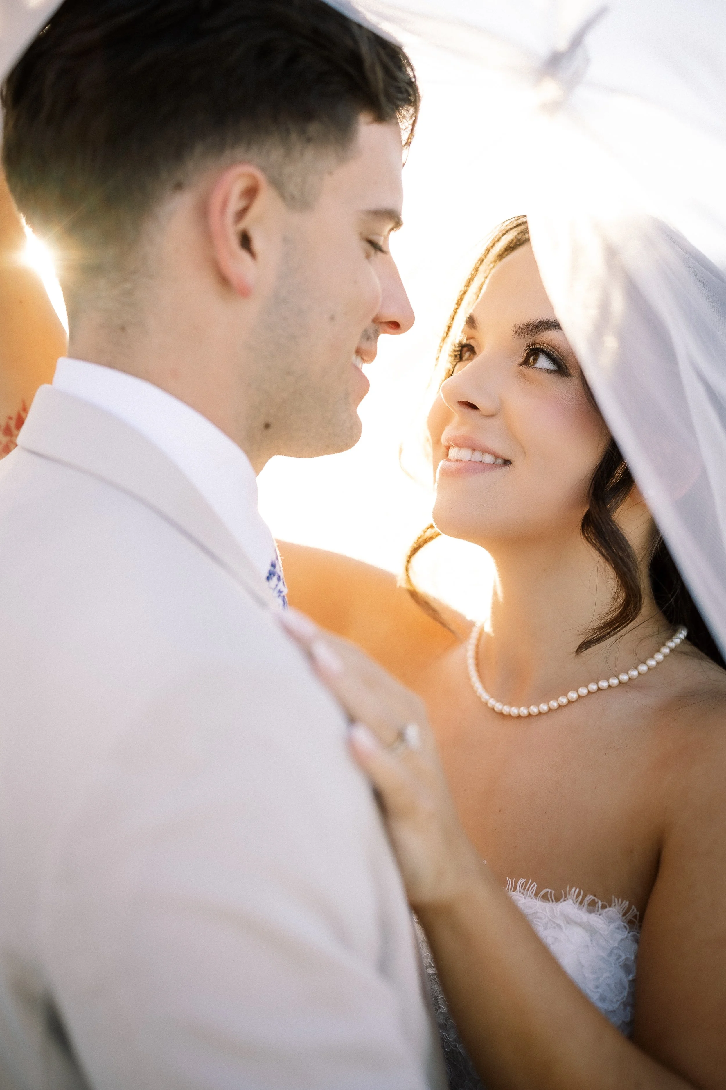 A bride and groom smiling at each other during their wedding, with sunlight in the background.