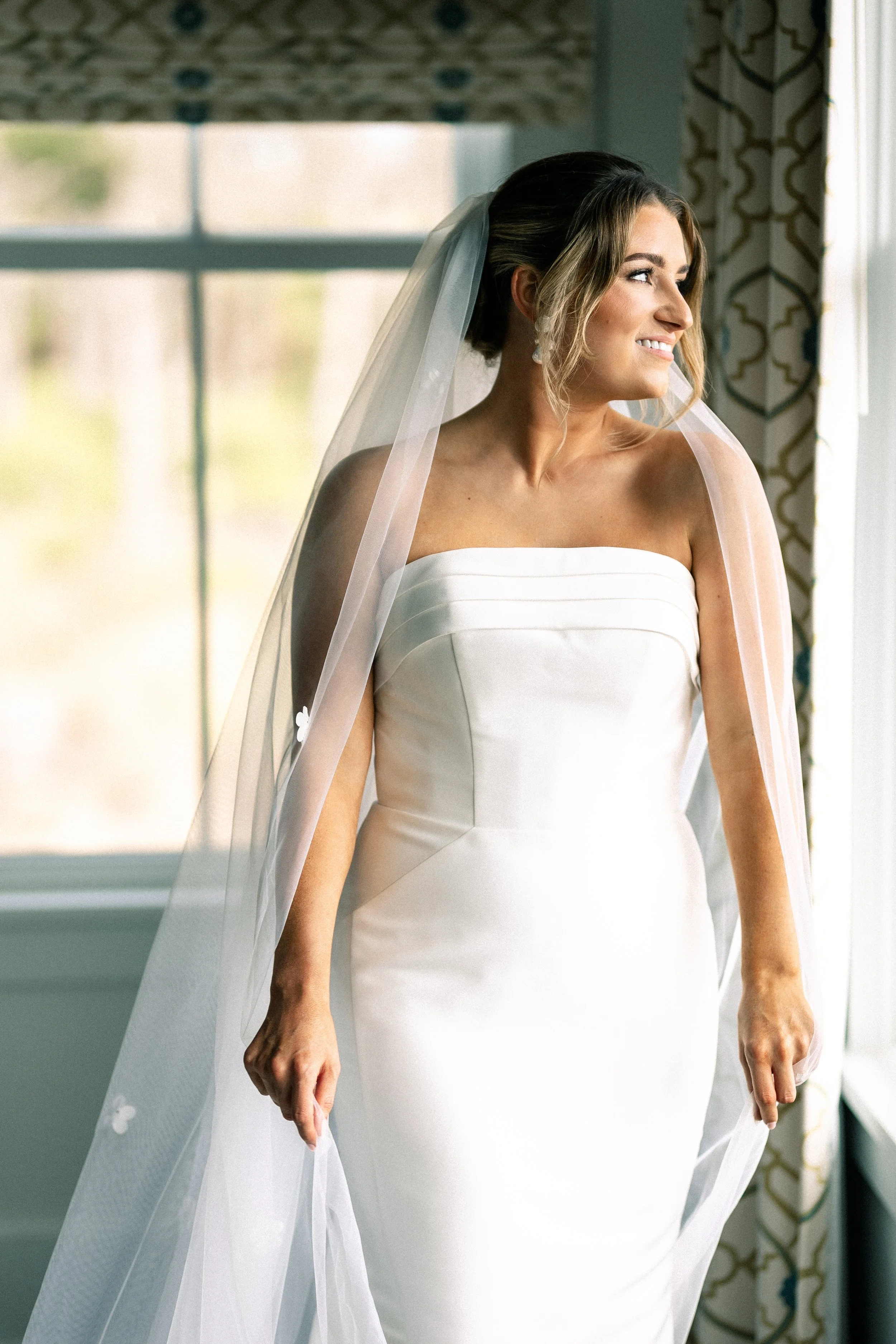 A bride in a strapless white wedding gown with a veil, smiling and looking out of a window.