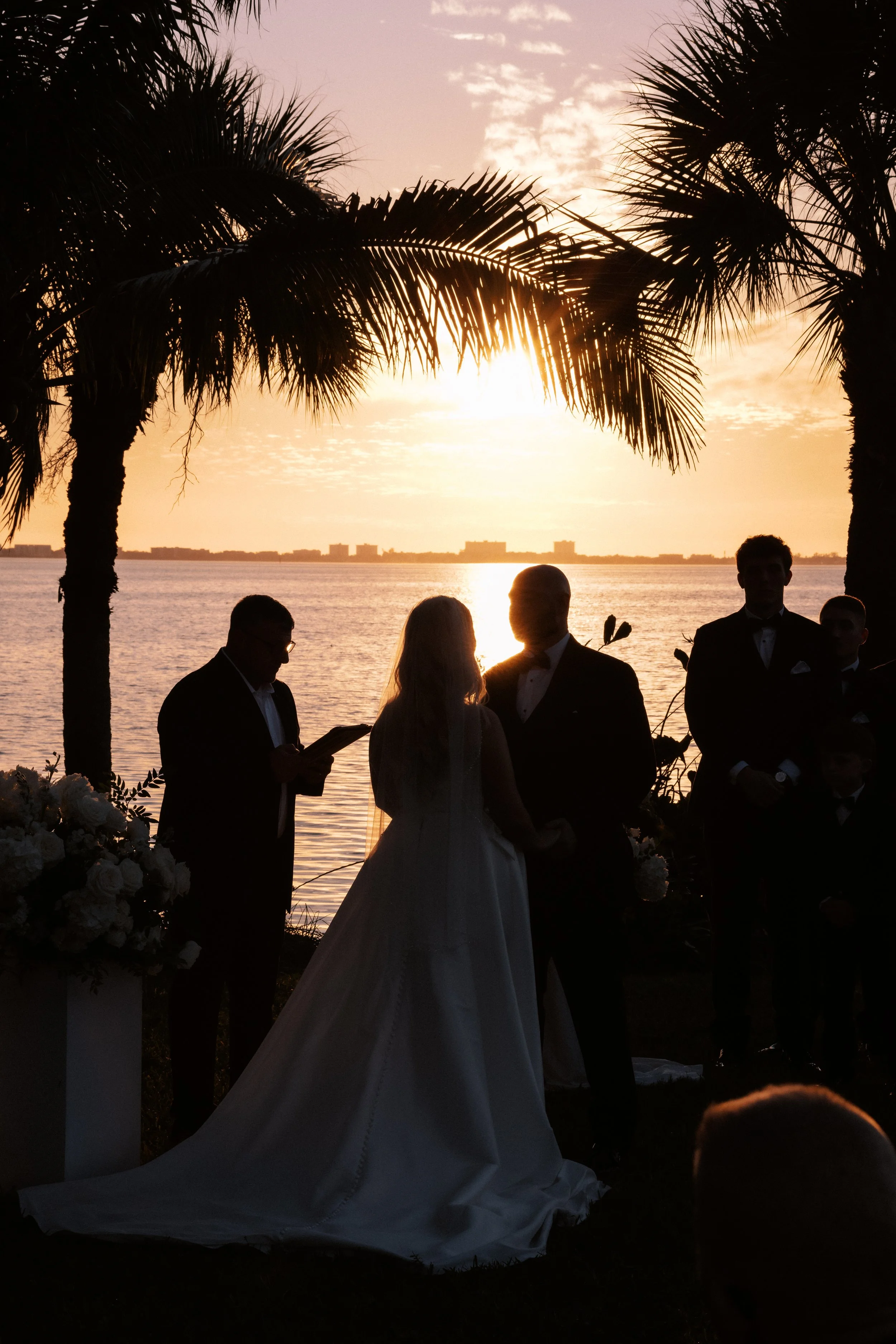 Silhouetted couple exchanging vows at sunset with palm trees and water in the background during a wedding ceremony.