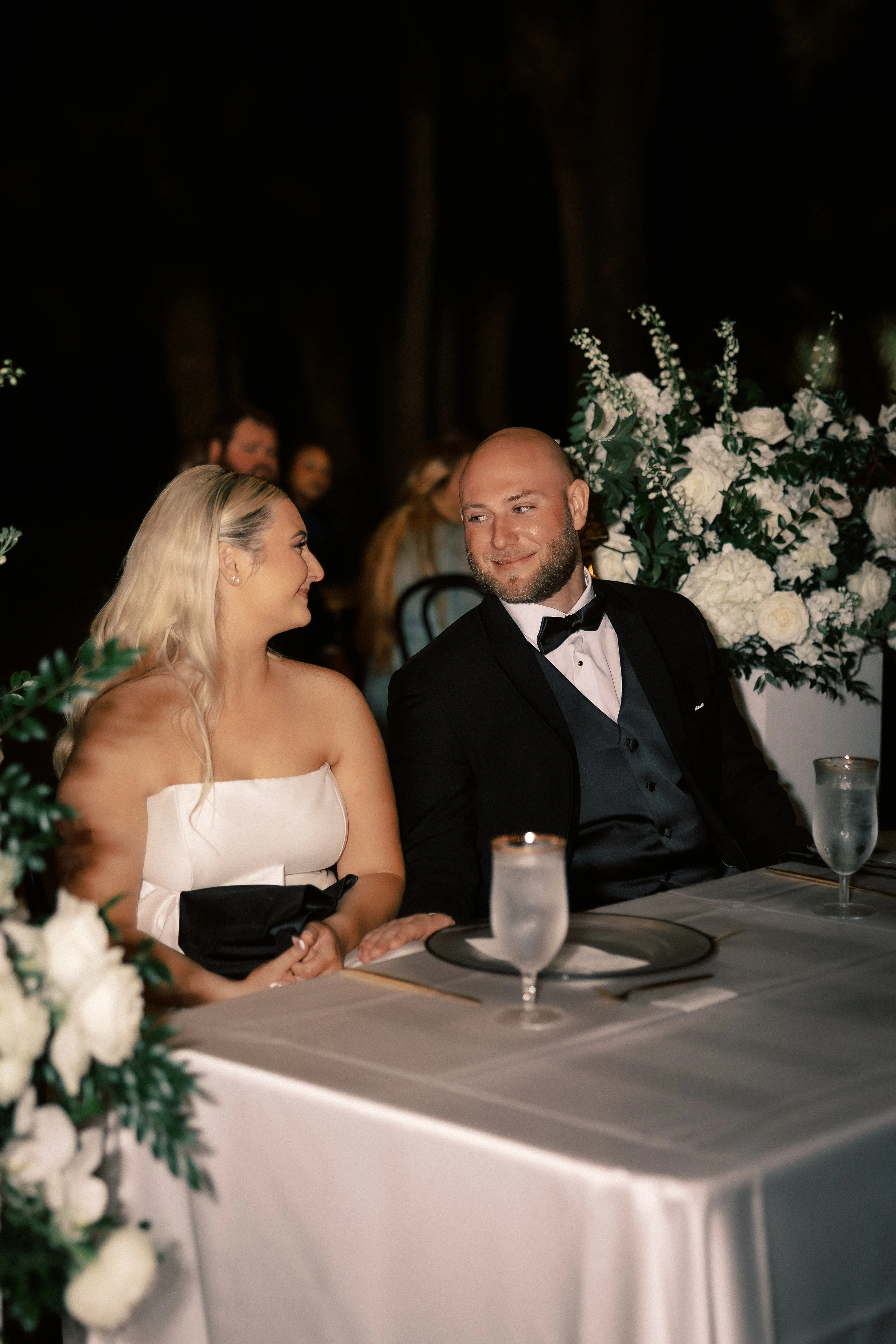 A couple dressed in formal wedding attire sits at a decorated table, smiling and looking at each other, with floral arrangements in the background.