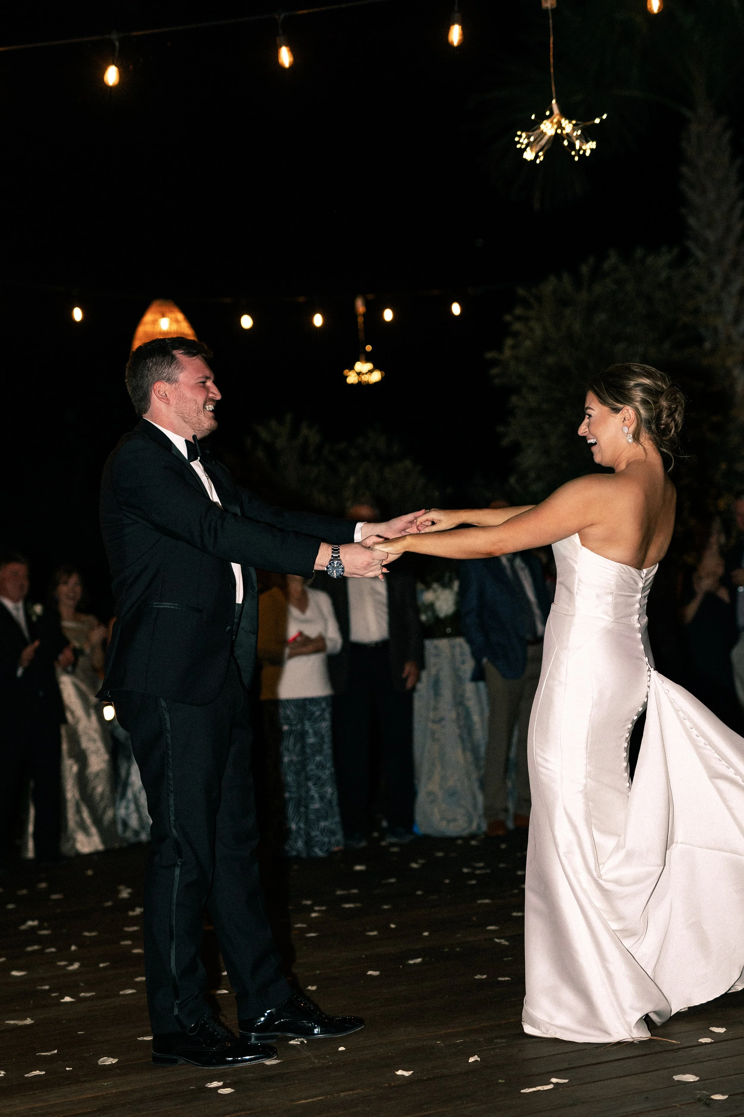 A bride and groom dancing at their wedding reception at night with string lights overhead and guests in the background.