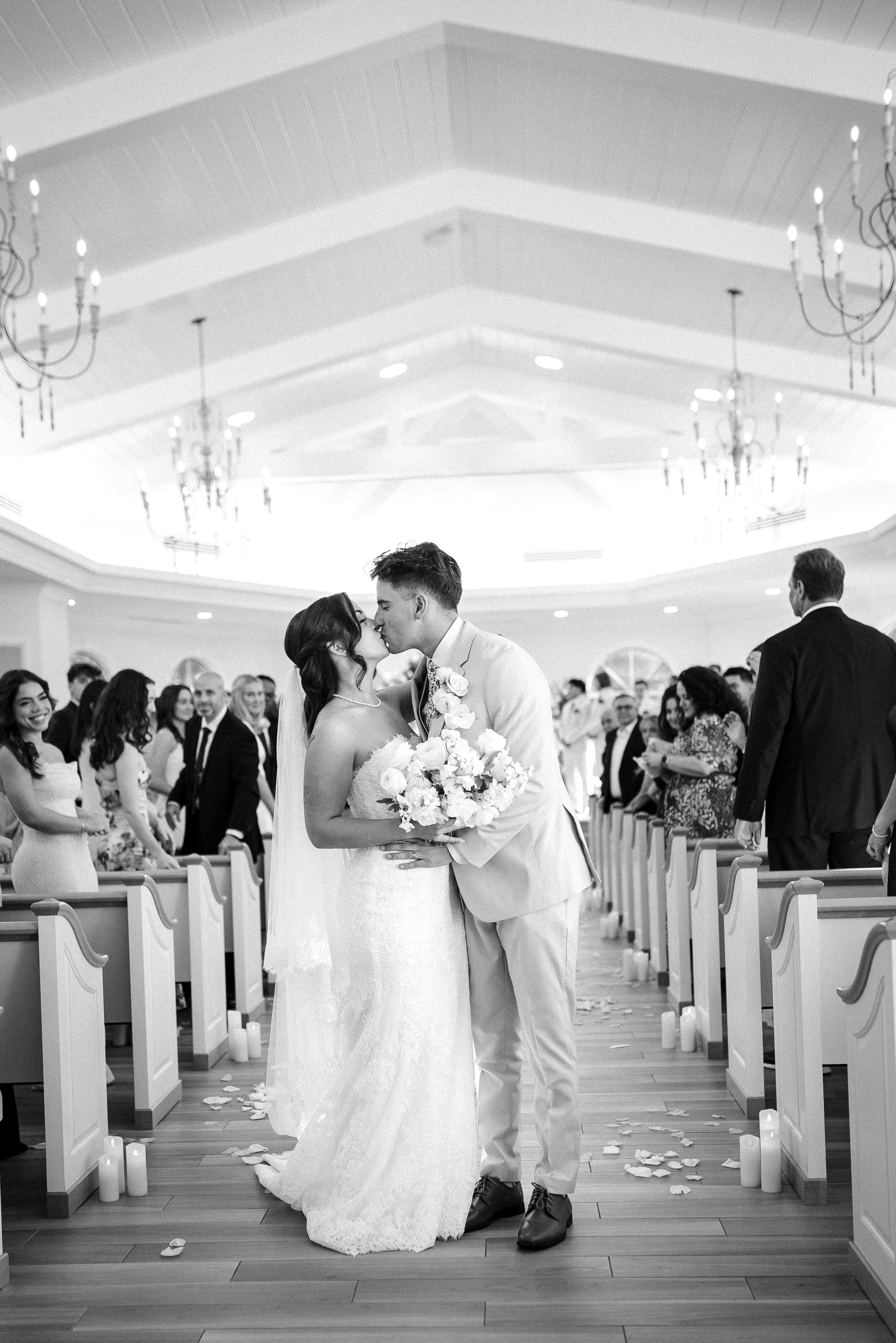 A bride and groom kiss at their wedding ceremony in a church filled with guests in the background. The bride holds a bouquet of flowers, and the scene is in black and white.