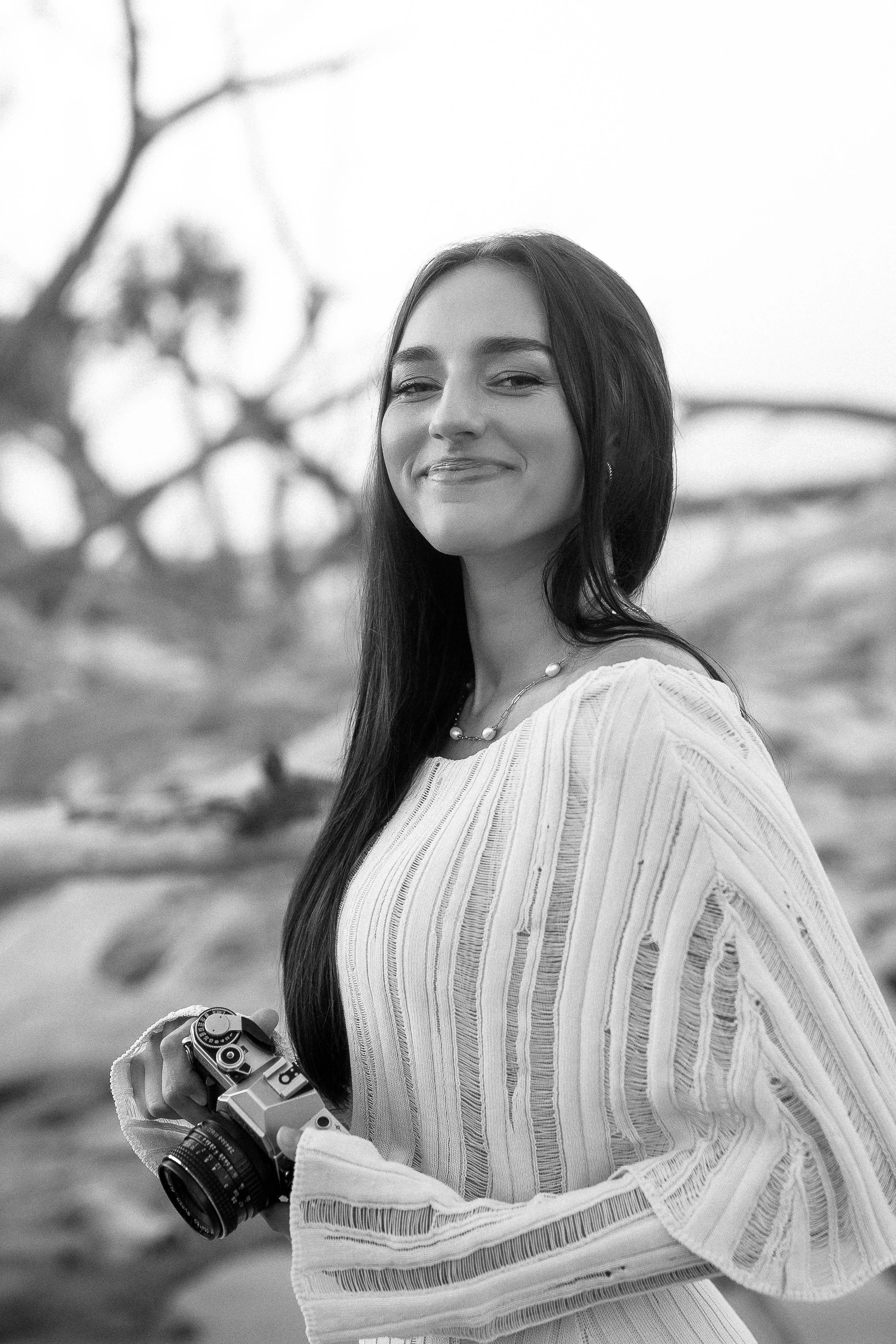 Black and white photo of a young woman with long dark hair, smiling, holding a vintage camera, dressed in a striped blouse or dress, outdoors with a blurred natural background.