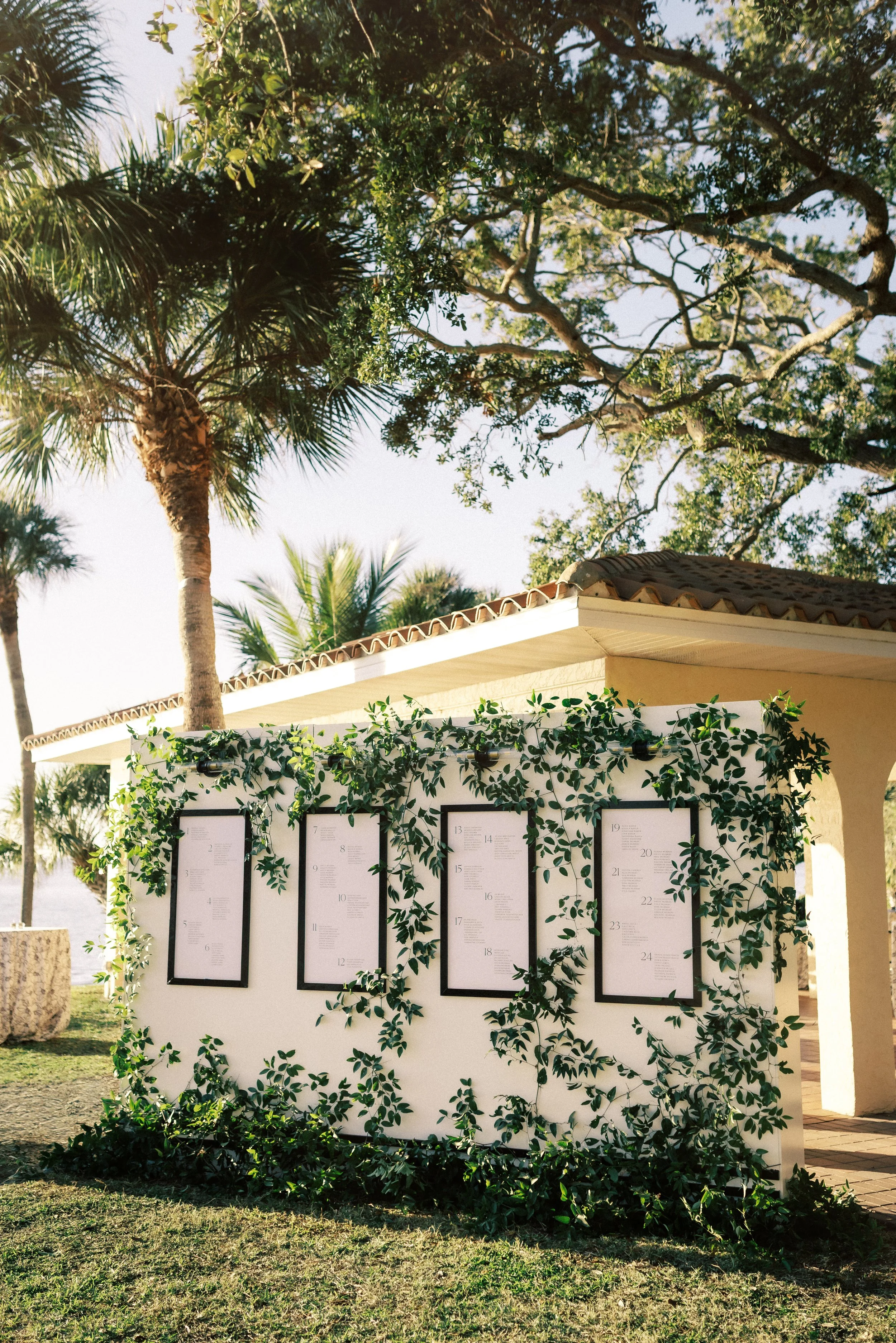 Outdoor scene with a white display board decorated with green foliage, containing multiple framed seating charts or event programs. There are tall palm trees and a building with a tiled roof in the background, under a clear sky.