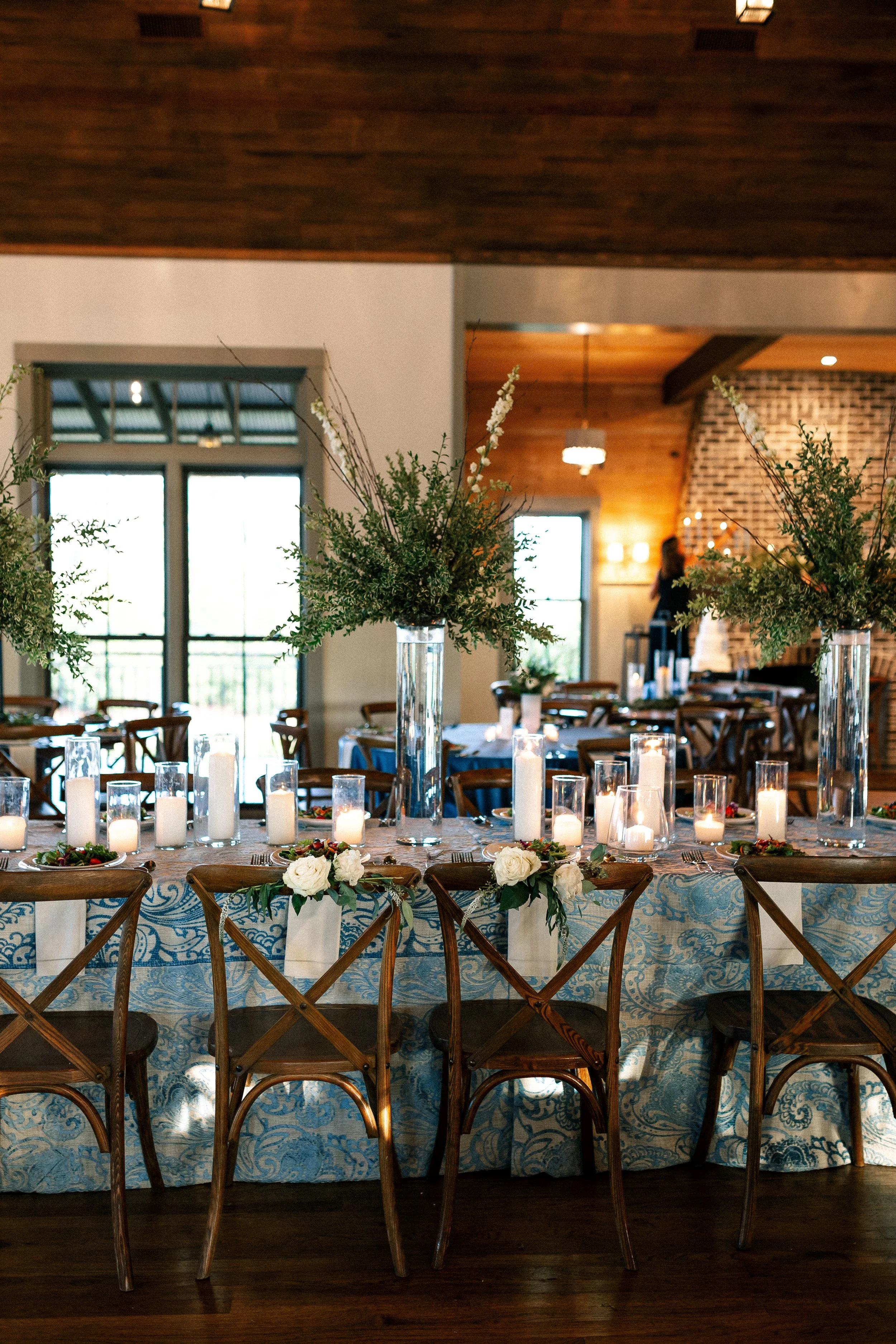 Decorated dining table with white roses, candles, and greenery in tall vases at a wedding or event reception venue with wooden accents and a brick fireplace in the background.
