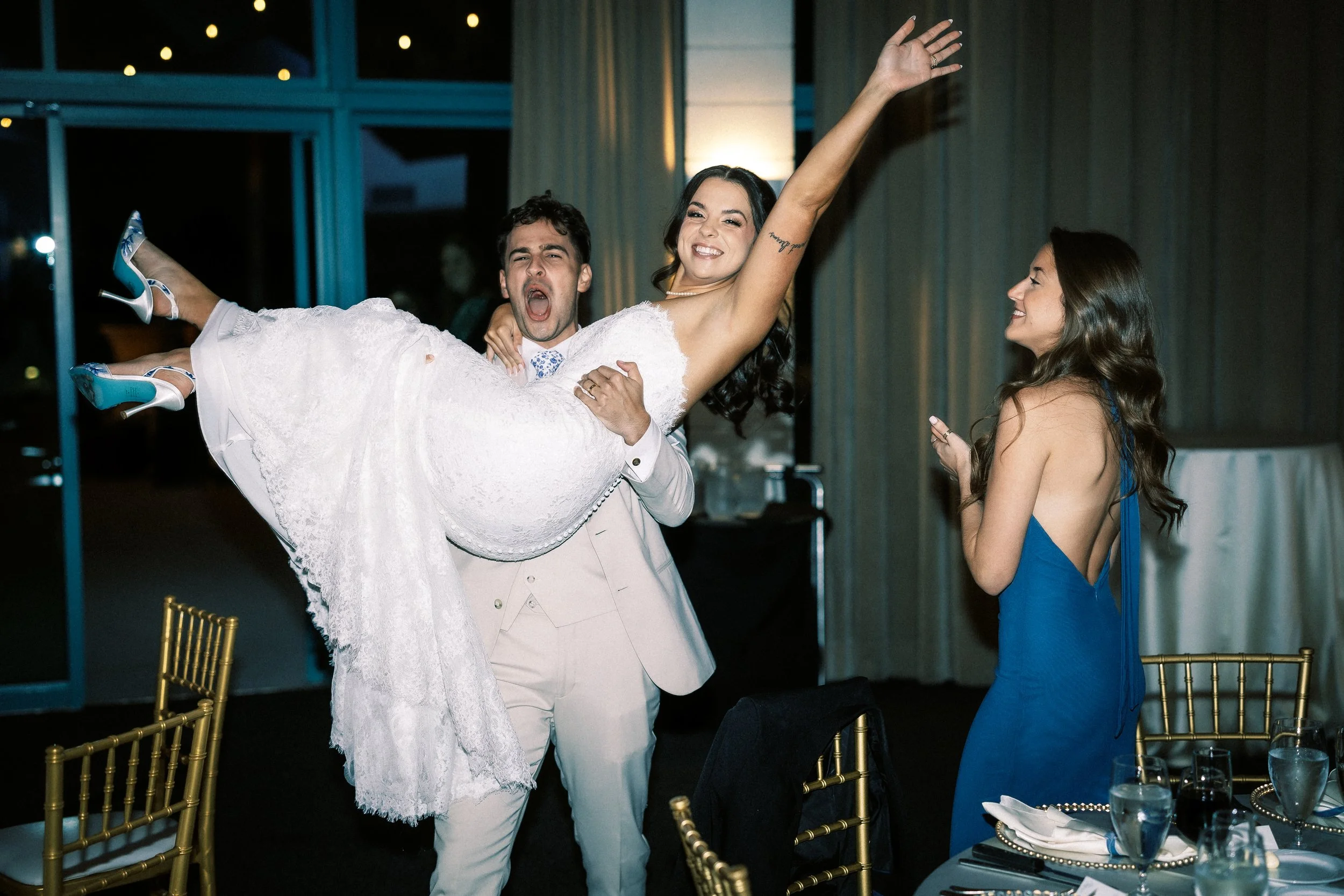A groom in a light suit is holding a bride in a white wedding dress, lifting her and dancing at a wedding reception. The bride is smiling with her arm raised, and another woman in a blue dress stands nearby, clapping and smiling.