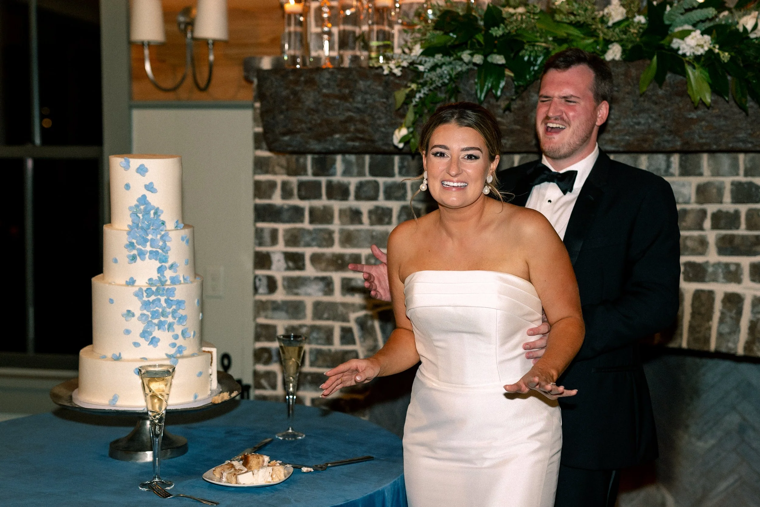 A bride and groom at their wedding reception, standing next to a wedding cake with blue flowers, champagne glasses, and a plate of cake on a table with a blue tablecloth, in front of a brick fireplace with floral decorations.
