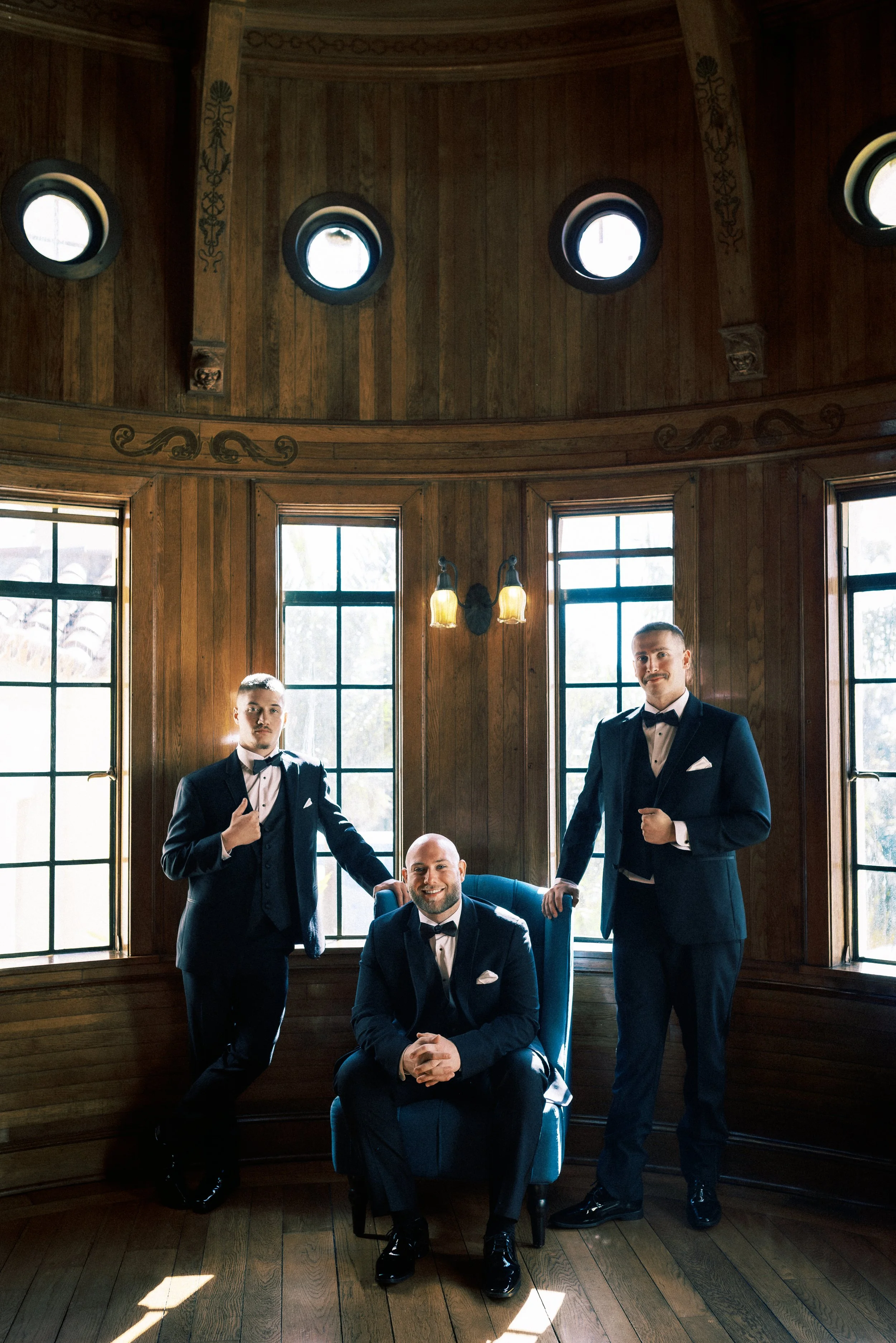 Three men dressed in black tuxedos with bow ties in a wood-paneled room with large windows and decorative lighting, one sitting in a blue armchair and two standing on either side.