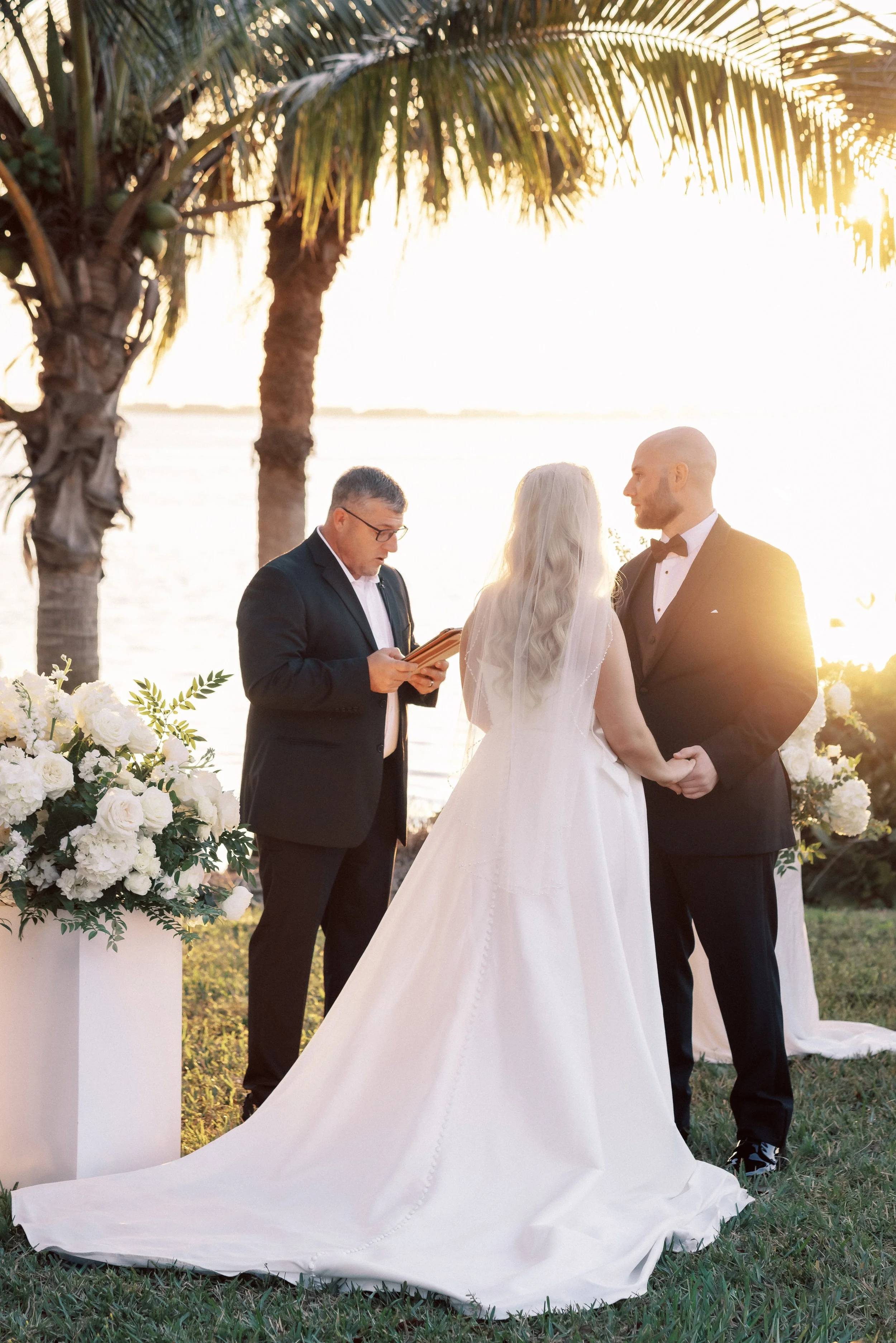 A couple getting married outdoors by a body of water during sunset, with a person officiating, surrounded by white flowers and palm trees.
