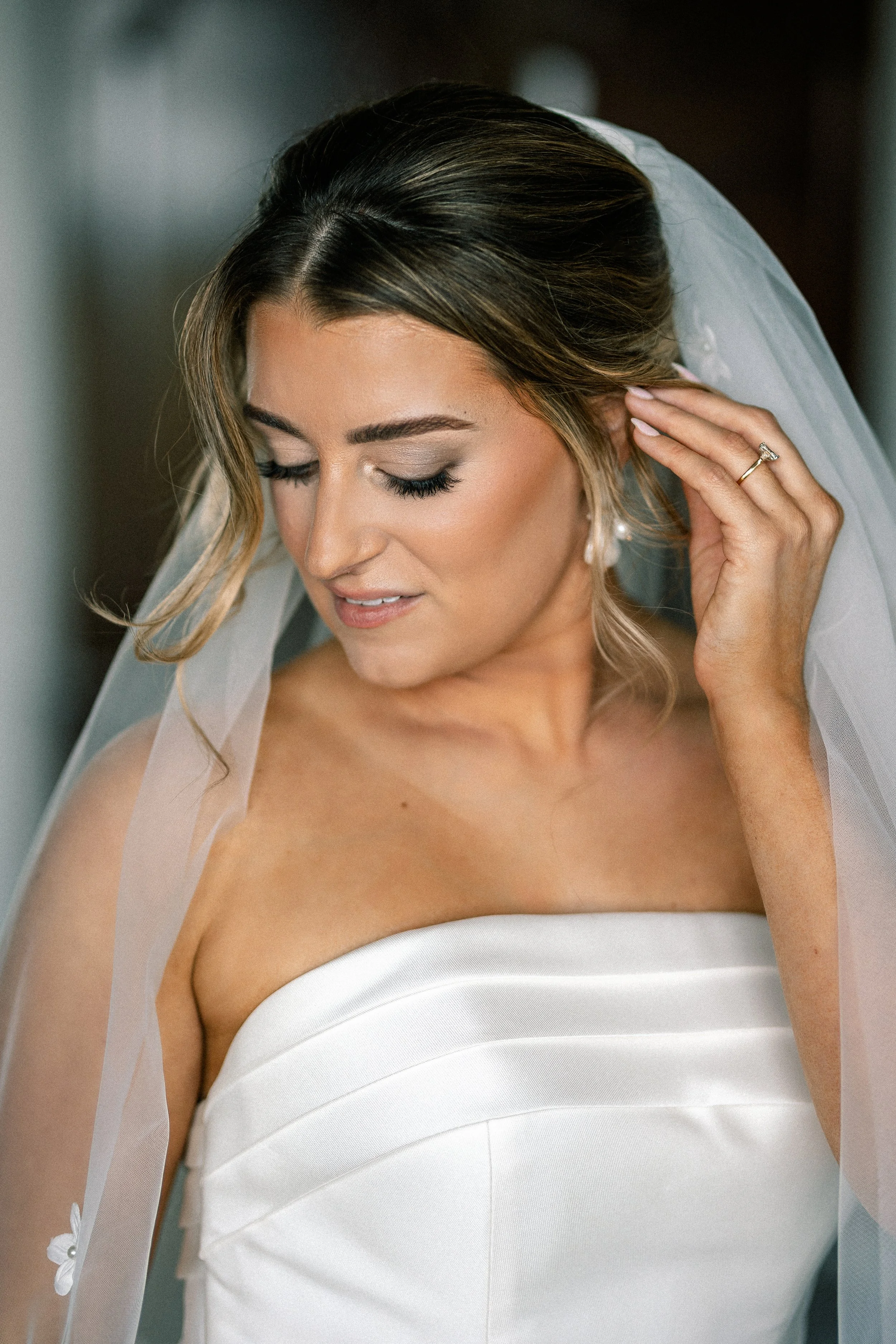Close-up of a bride in a strapless white wedding gown with a veil, touching her hair, with makeup and a wedding ring visible.