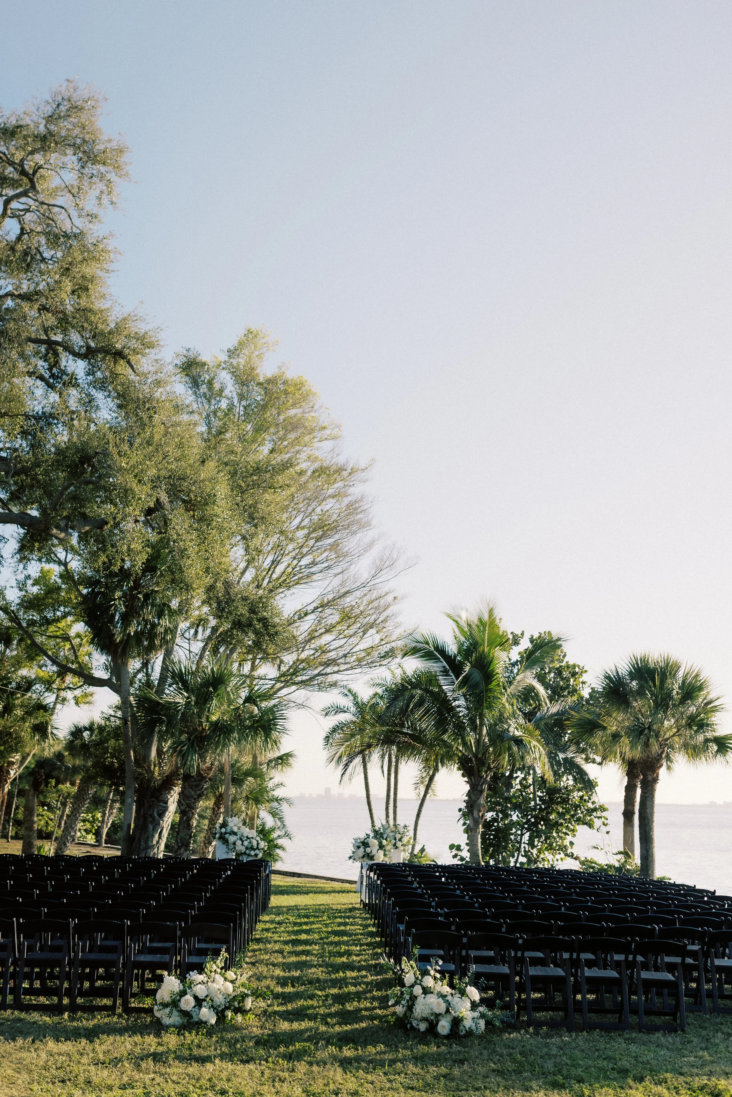 Outdoor wedding ceremony setup with rows of black chairs on a grassy area, decorated with white floral arrangements, near palm trees by the water under a clear sky.
