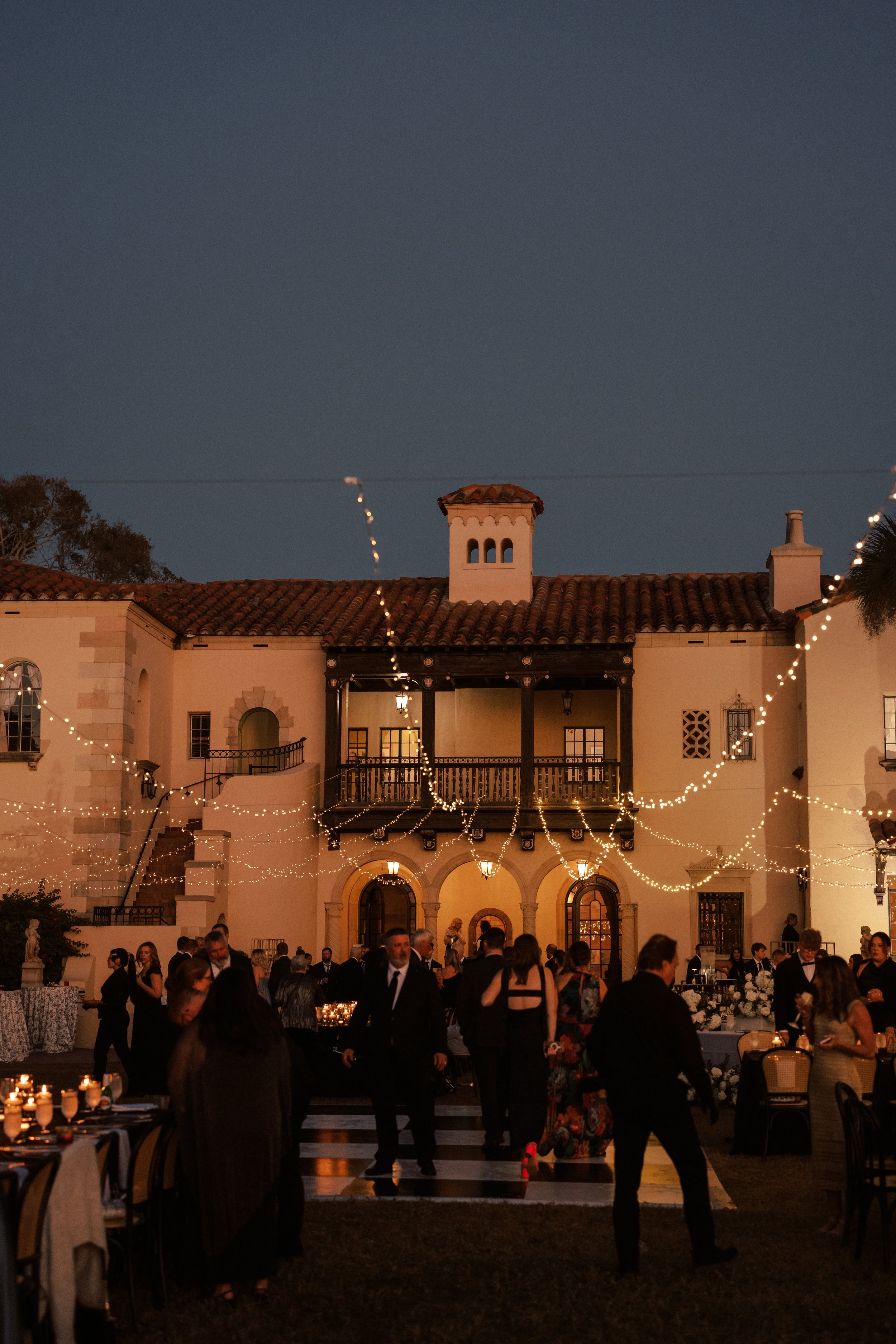 People attending a formal outdoor evening event with string lights, tables, and a large Mediterranean-style building in the background.