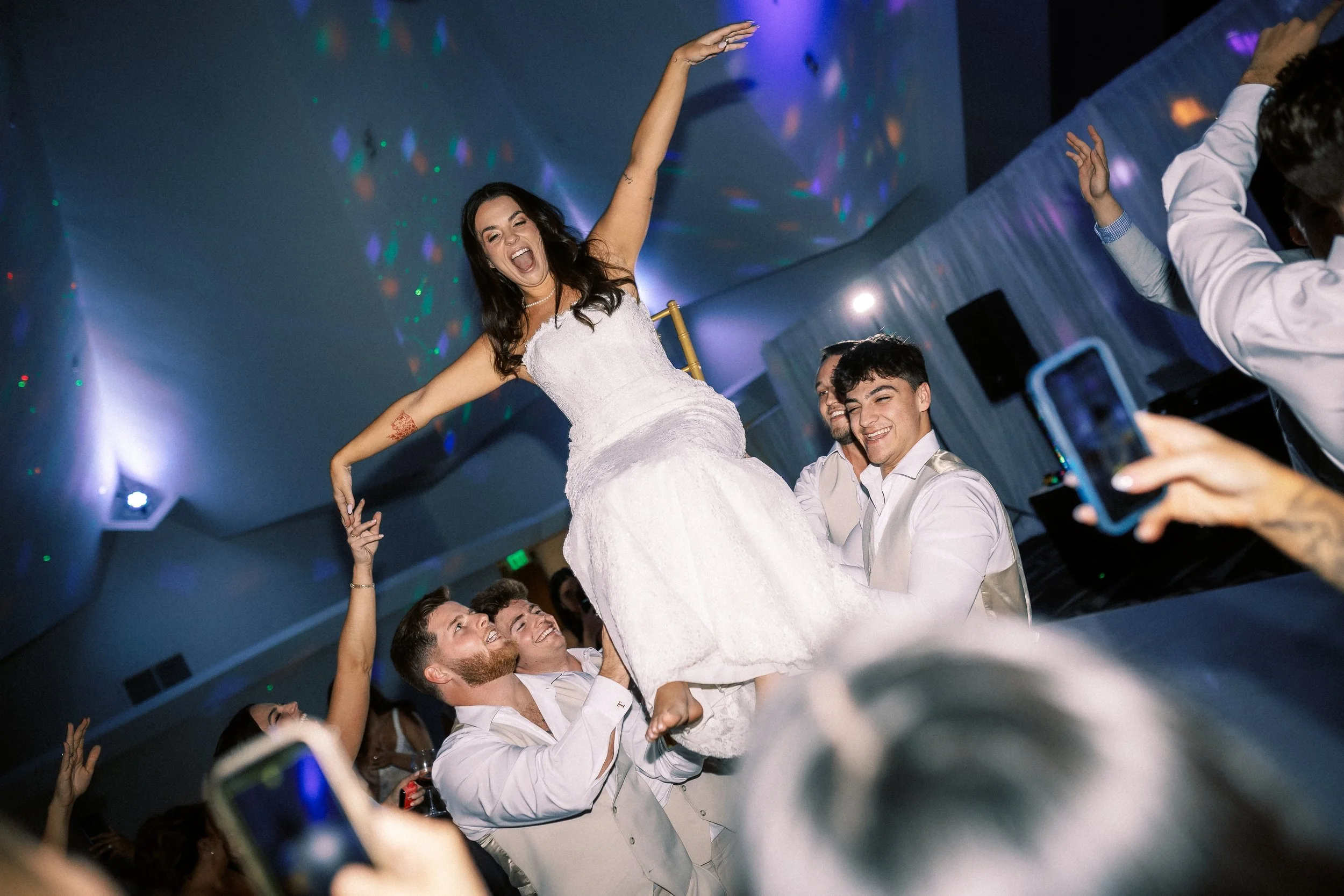 People celebrating at a wedding reception, with a woman in a white dress being lifted on the shoulders of others in a decorated hall.