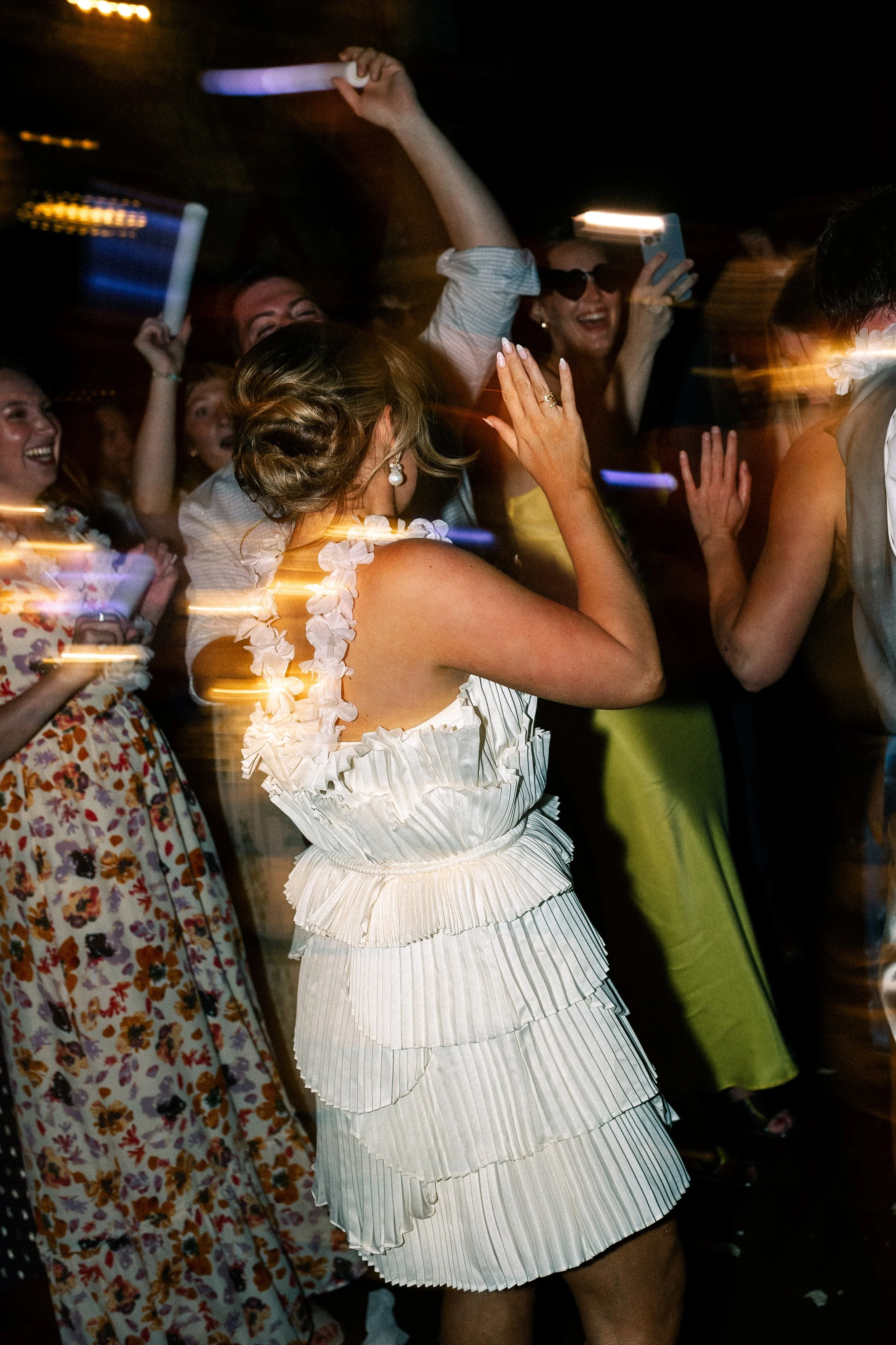 People dancing and smiling at a celebration or party, with women in colorful dresses and one woman in a white ruffled dress in the foreground.