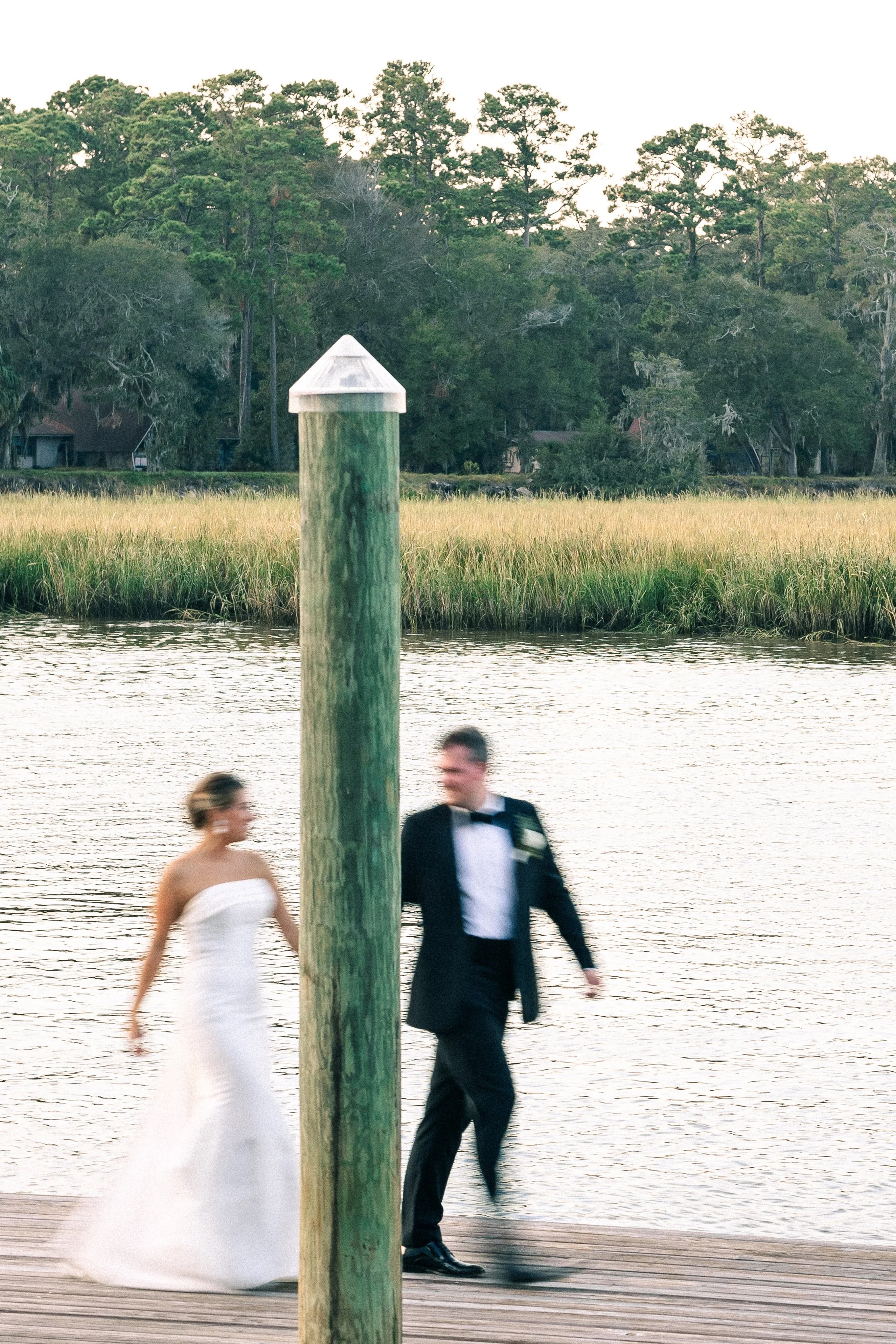 A blurred bride in a white wedding dress and groom in a black tuxedo walking on a wooden dock near water, with greenery and trees in the background.