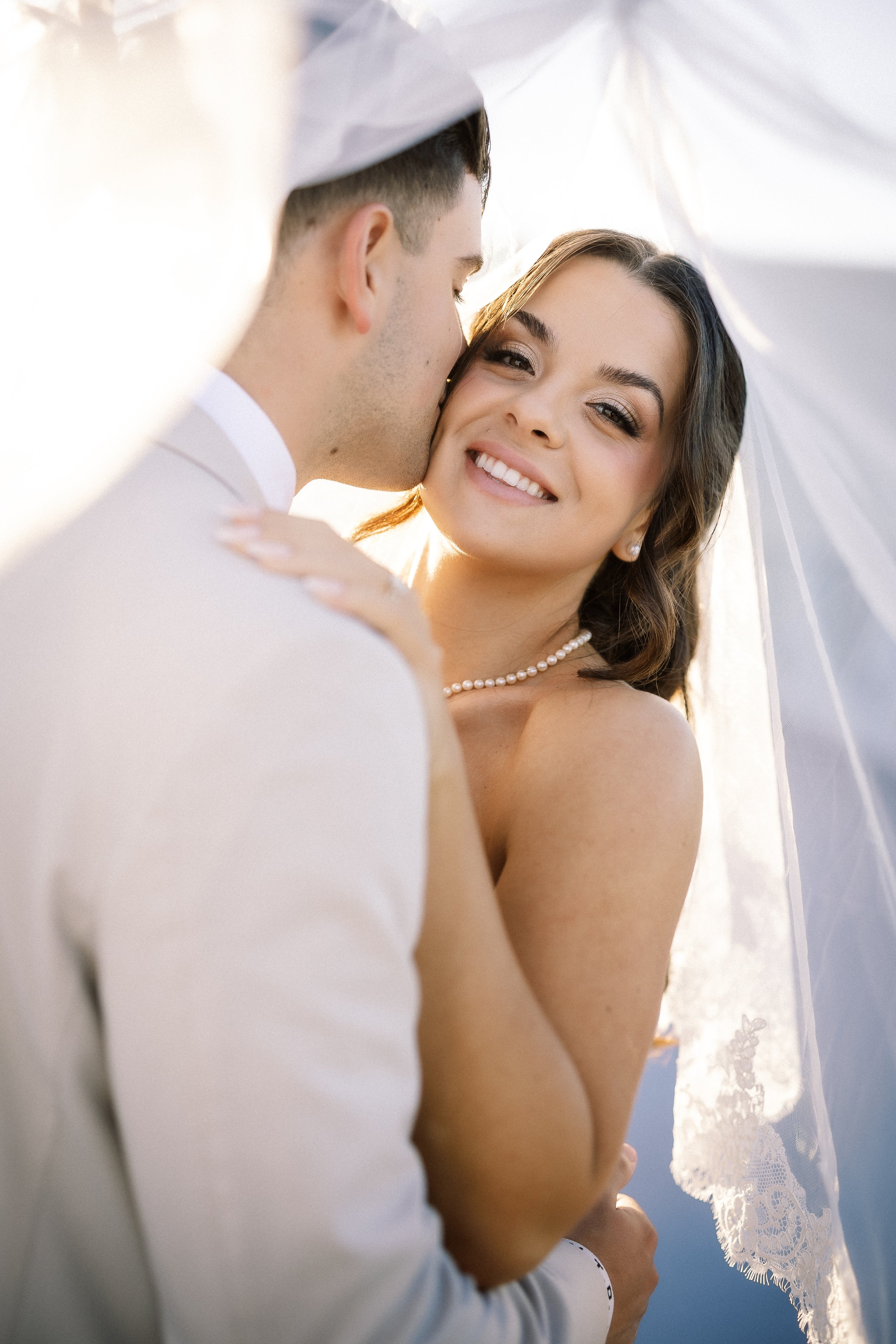 A bride and groom share a romantic moment under a white umbrella, with sunlight shining through, as the groom kisses the bride on her cheek. The bride has dark hair, a pearl necklace, and is smiling.