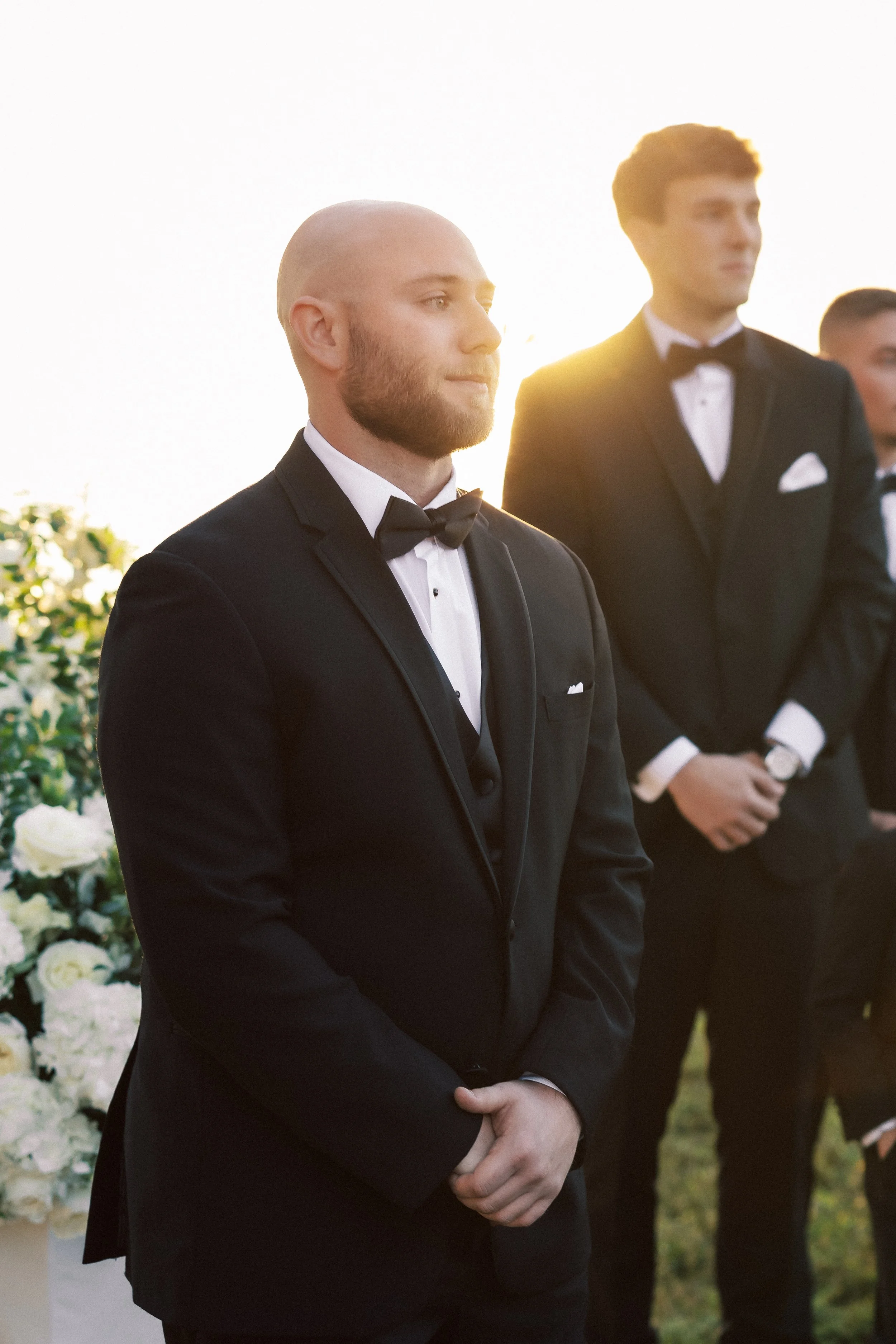 A man in a black tuxedo with a bow tie and white shirt standing outdoors during sunset, with two other men dressed similarly in the background, near a floral arrangement.