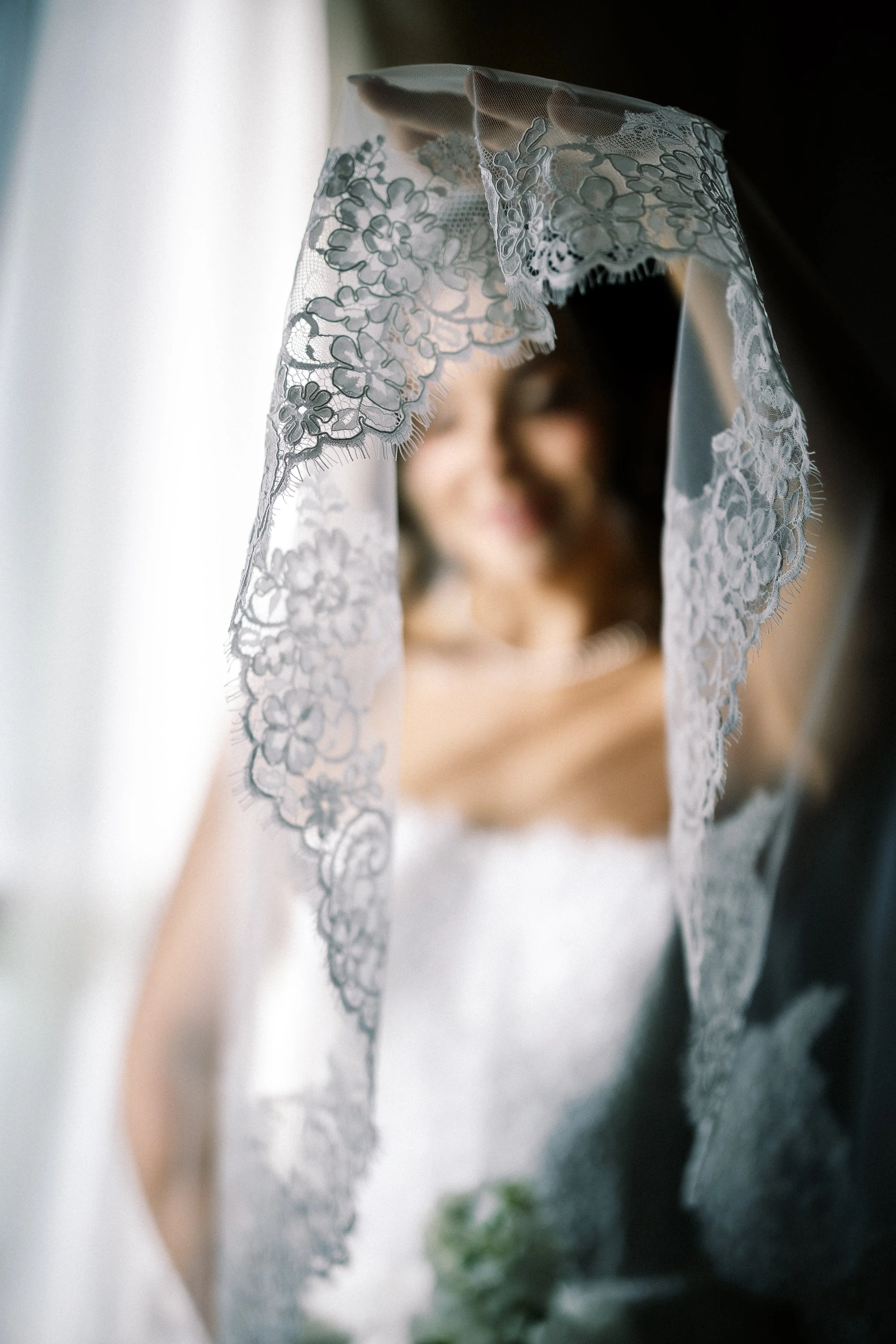 Close-up of a bride's veil with intricate floral lace, slightly out of focus, as the bride smiles in the background.