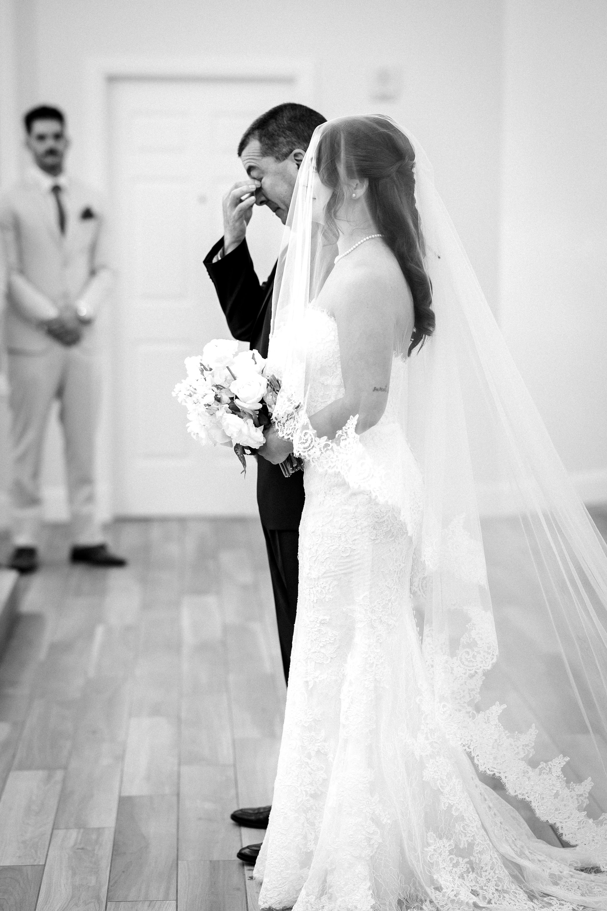 Black and white photo of a bride and groom during their wedding ceremony, with the groom wiping tears and the bride holding a bouquet of flowers.