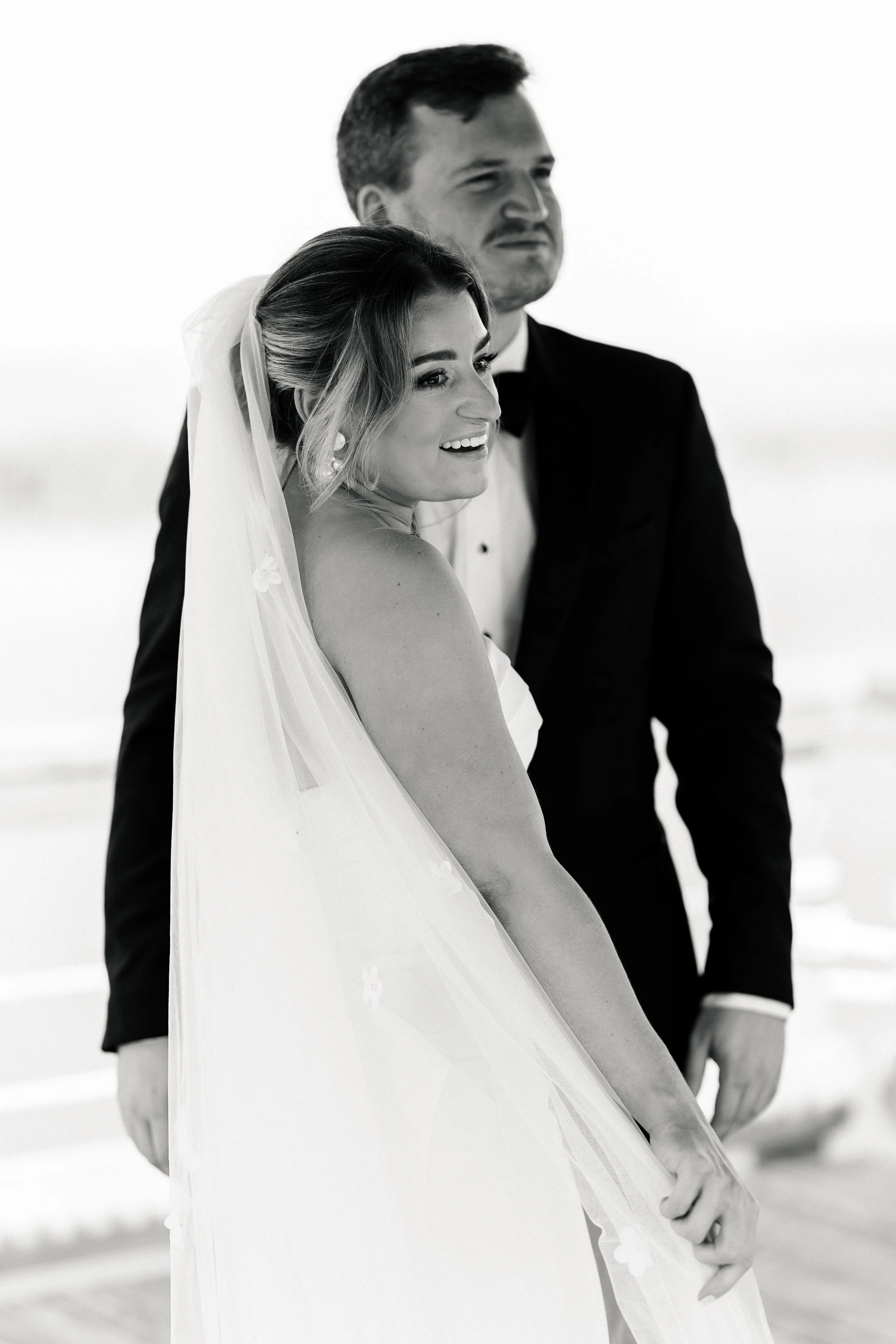 Black and white photo of a bride in a wedding dress with a veil and a groom in a tuxedo, standing outdoors.
