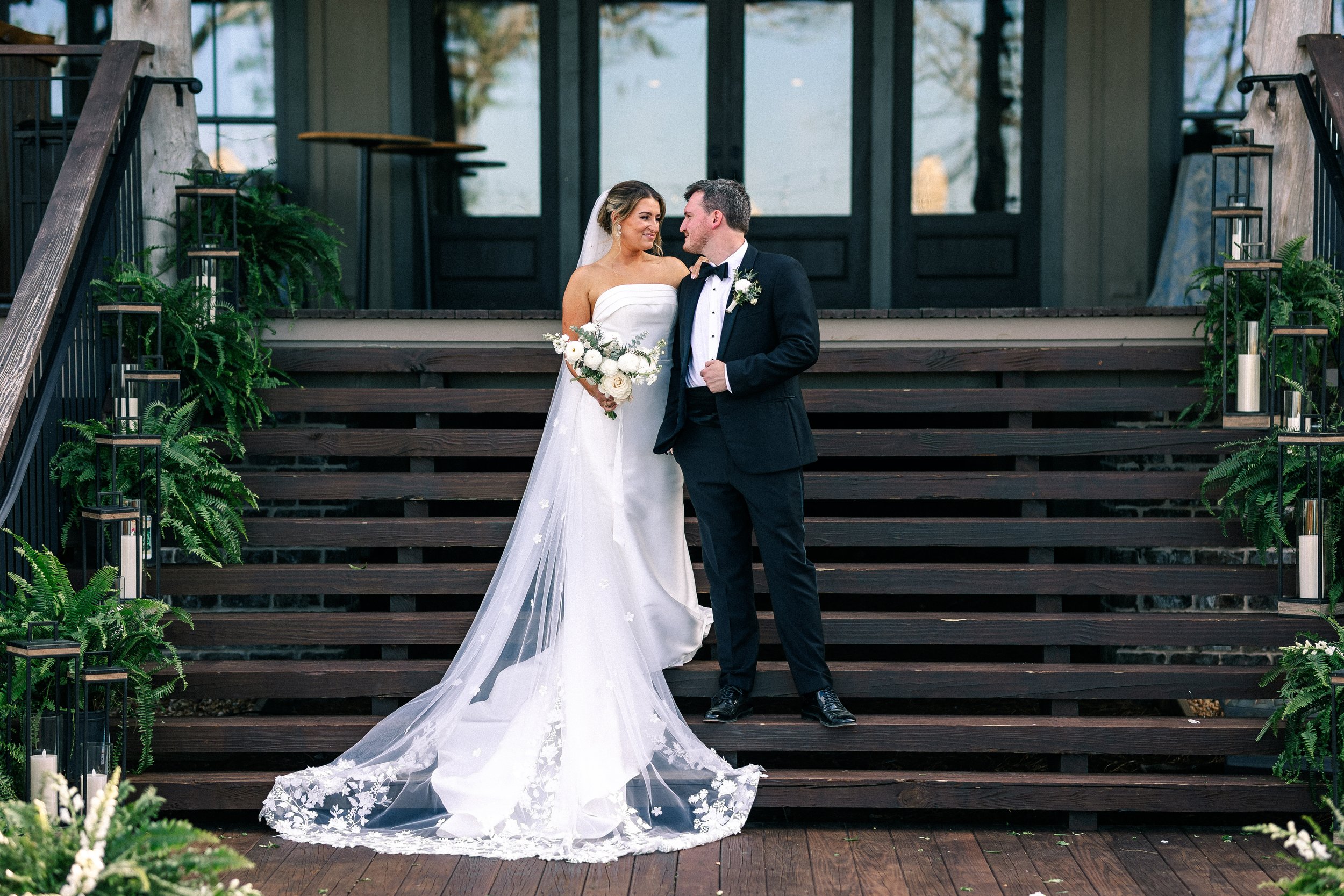 A bride and groom standing on outdoor stairs, looking at each other, with the bride holding a bouquet of white flowers, in front of a building with glass doors.