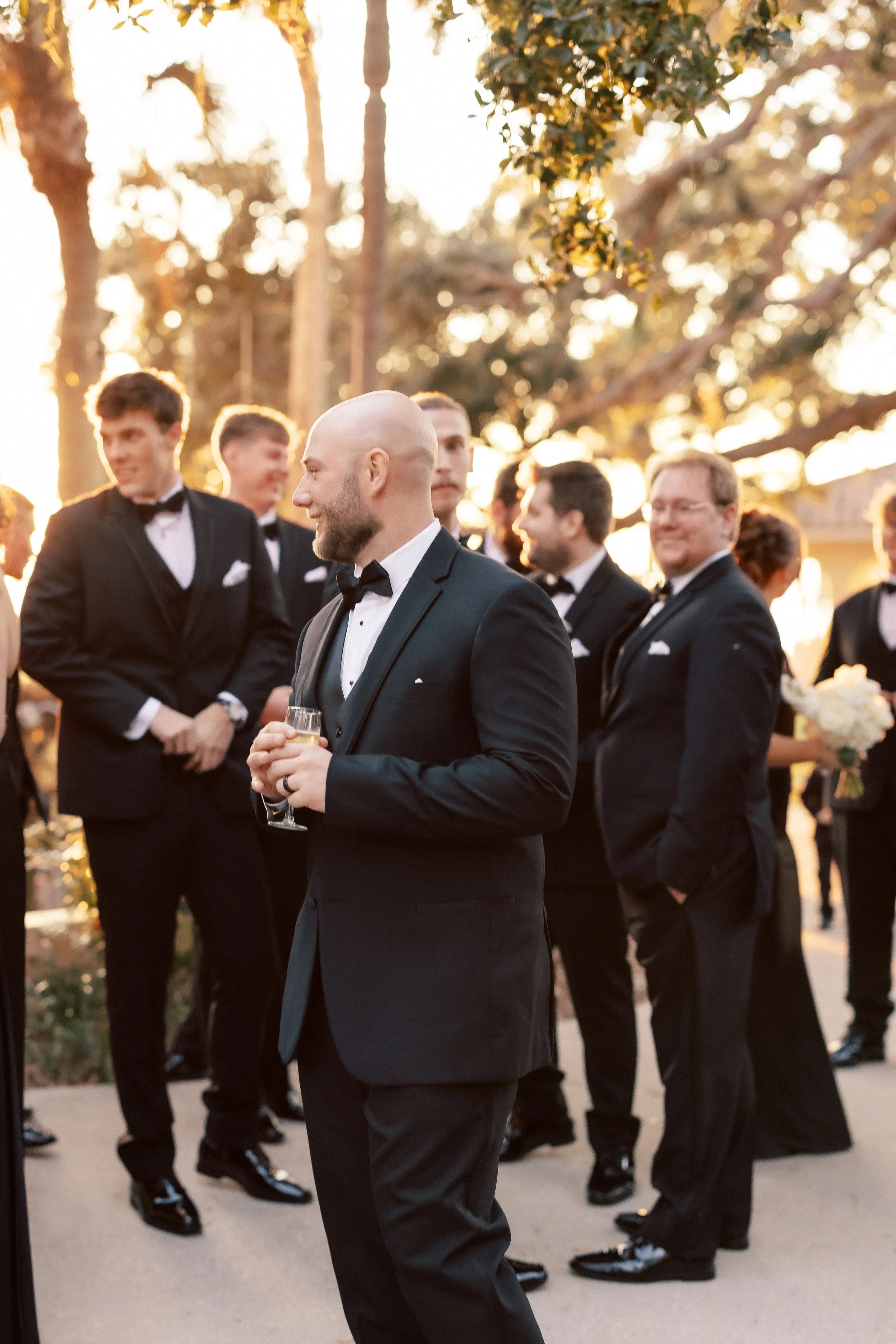 Groom and groomsmen at a wedding reception outdoors during sunset, dressed in black tuxedos, with one man holding a champagne glass.