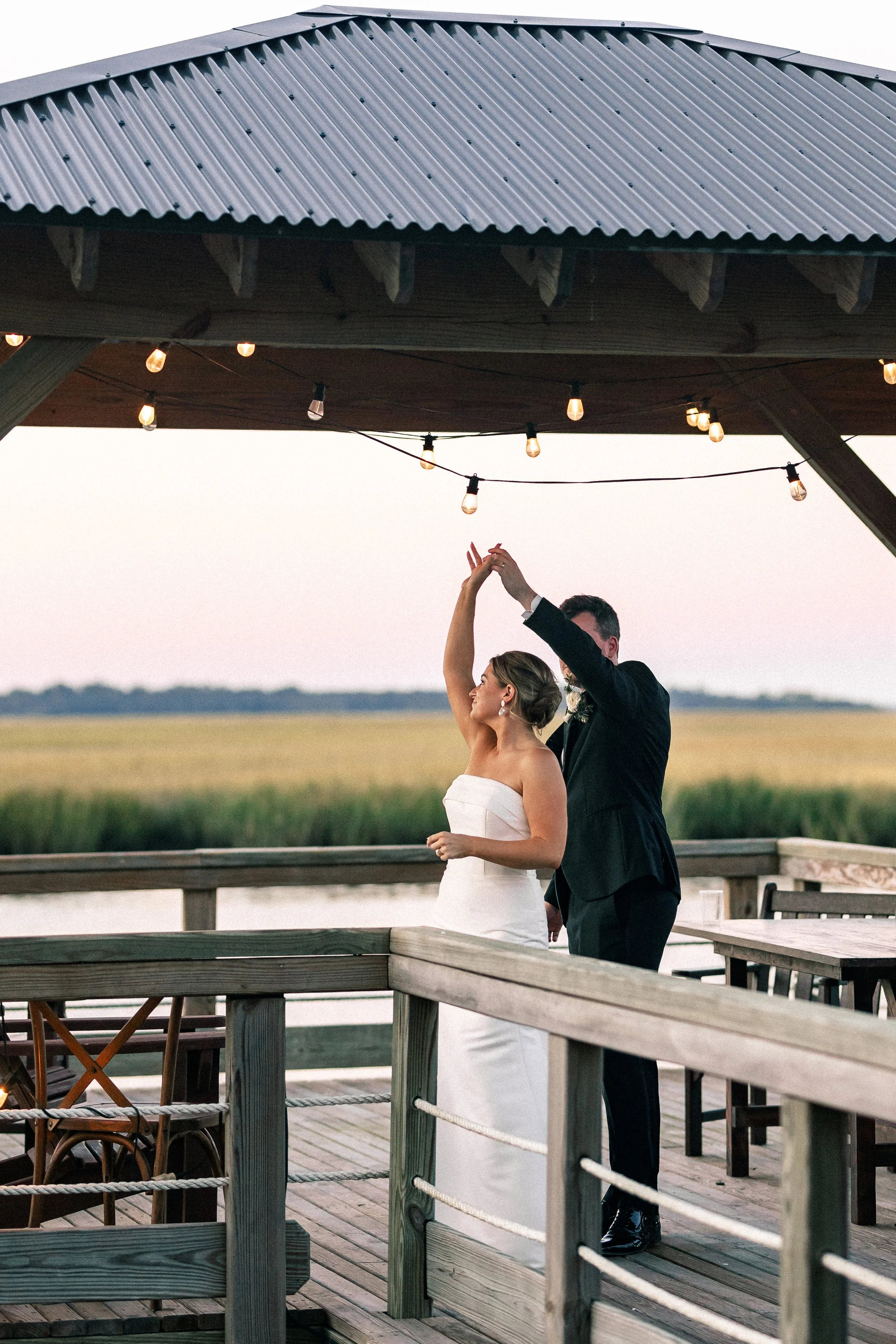 A bride and groom dancing on a wooden deck with a view of a marshland and a pink sky at sunset, under string lights.
