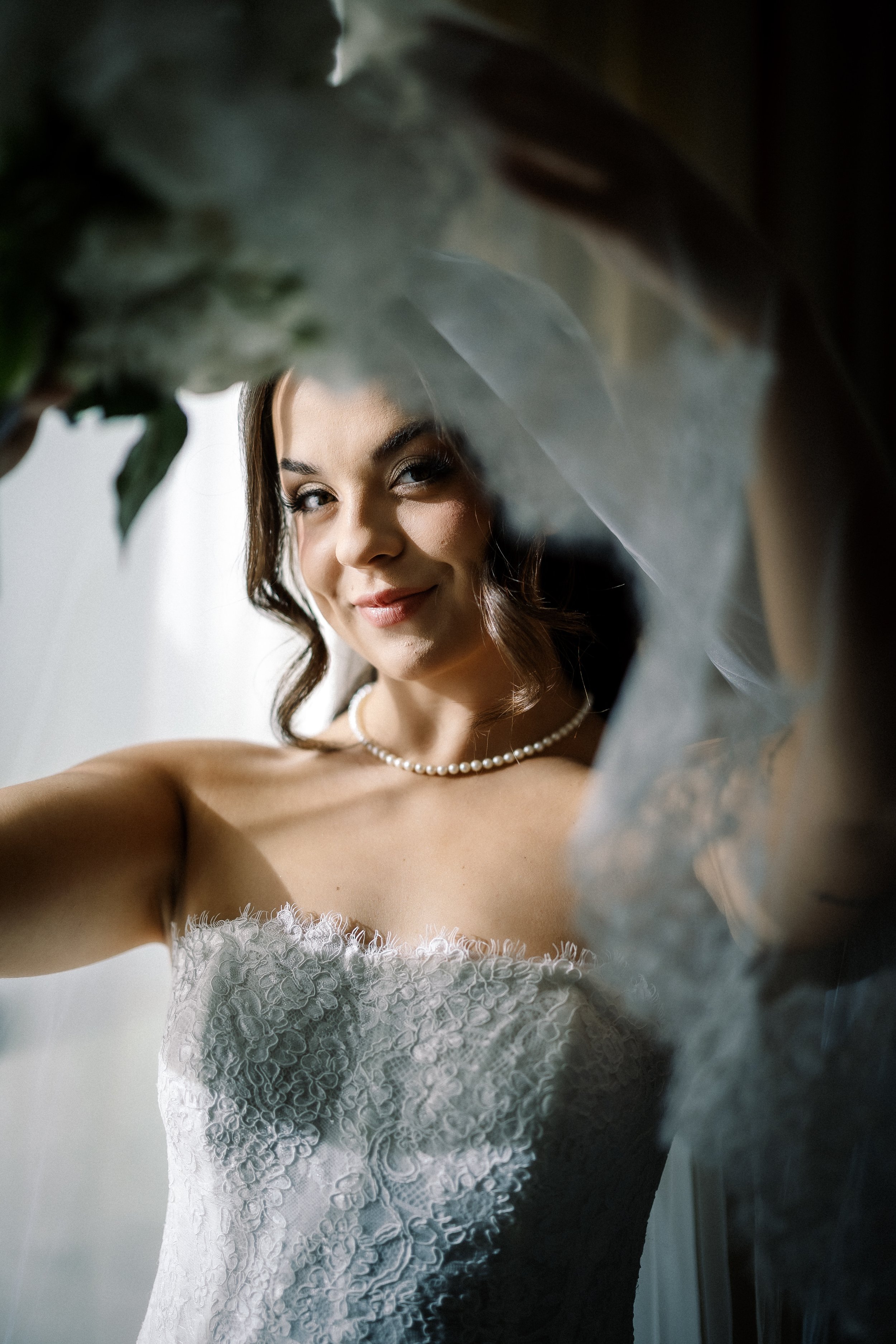 A bride in a lace wedding dress and pearl necklace taking a selfie, smiling through a camera lens with greenery and a curtain in the background.