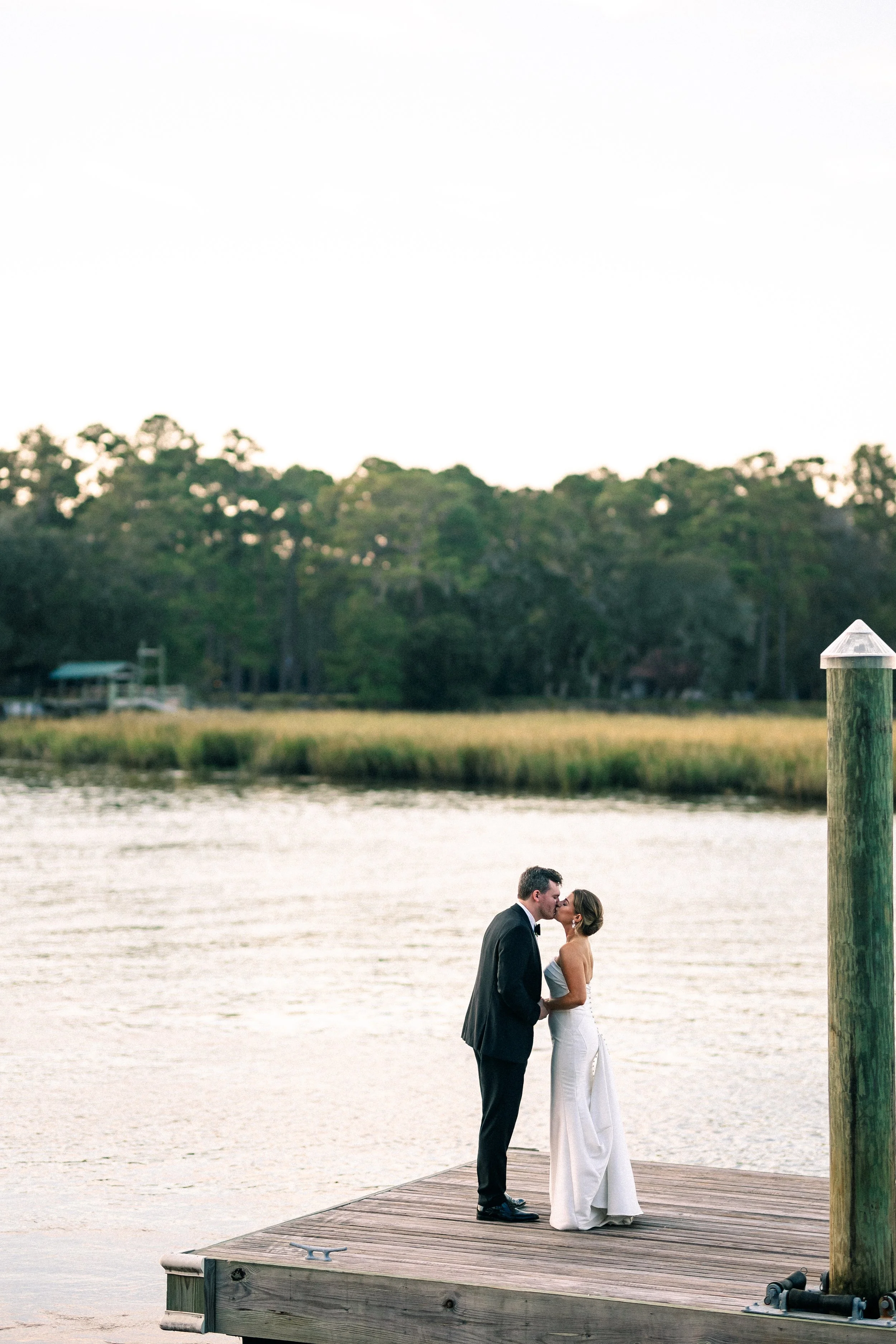 A bride and groom kissing on a wooden dock beside a body of water, with trees in the background during sunset