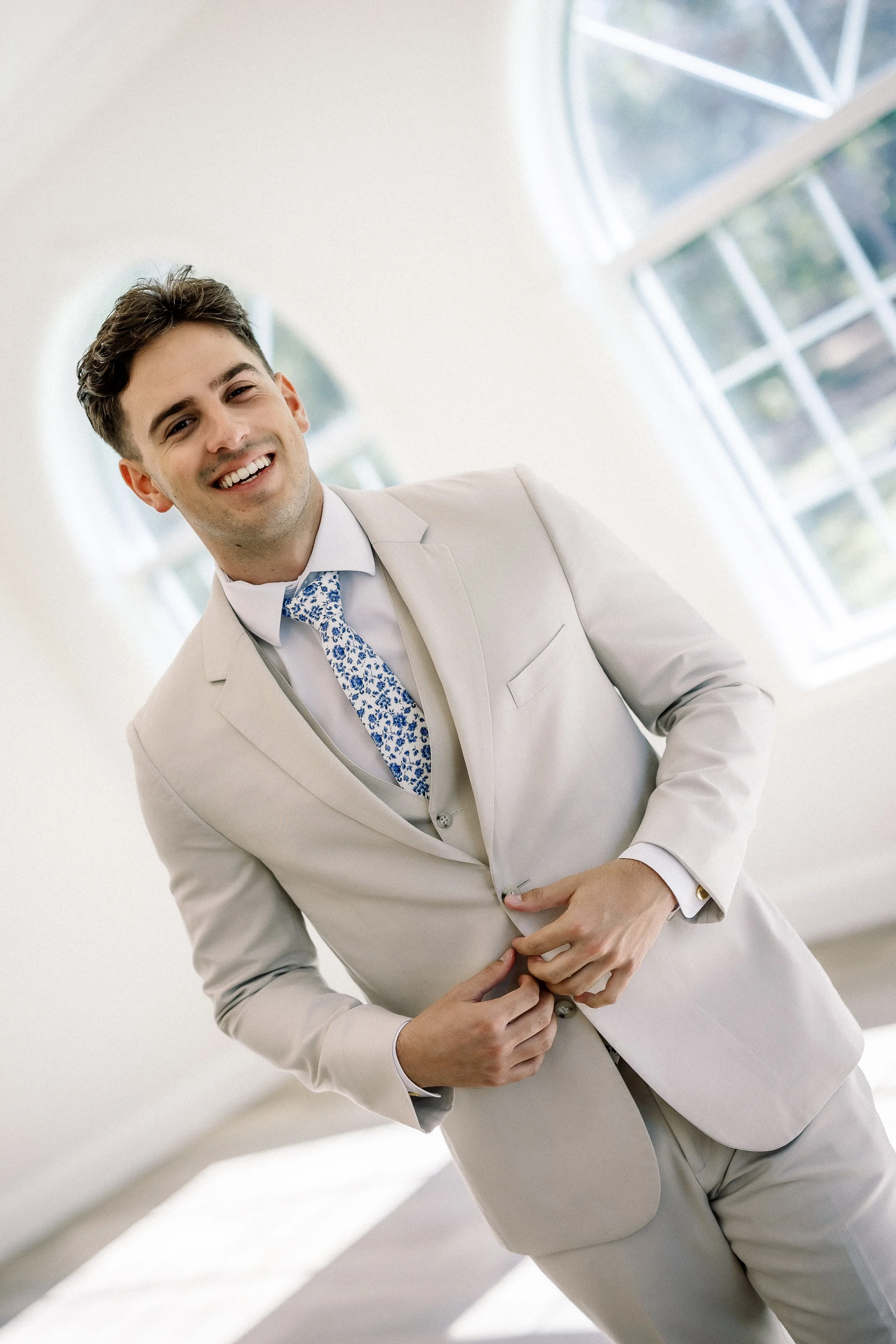 A young man in a light beige suit smiling, adjusting his jacket, standing in a bright room with large arched windows in the background.